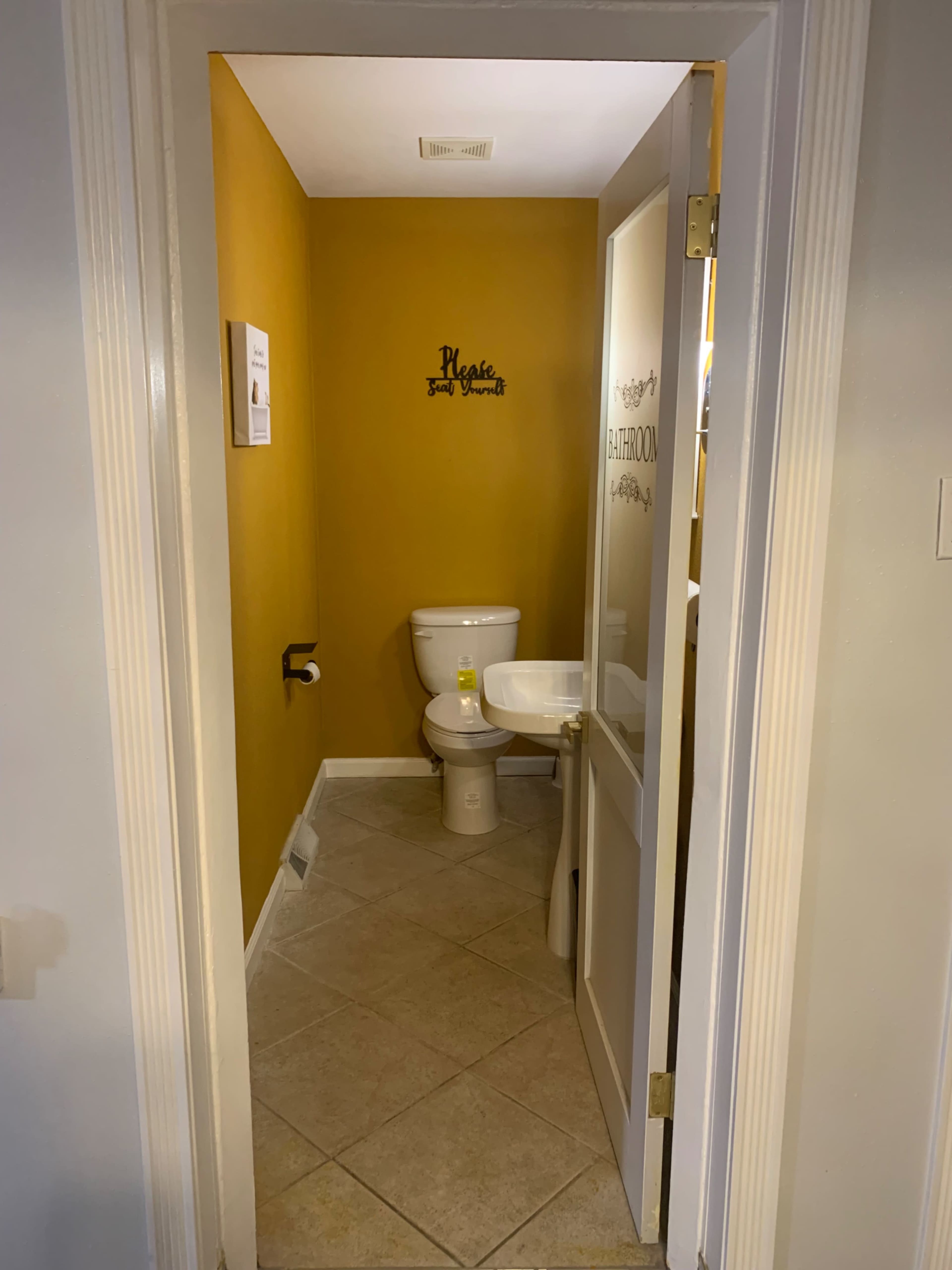 The image shows a small bathroom with yellow-painted walls, containing a toilet and a sink, viewed from an open door.