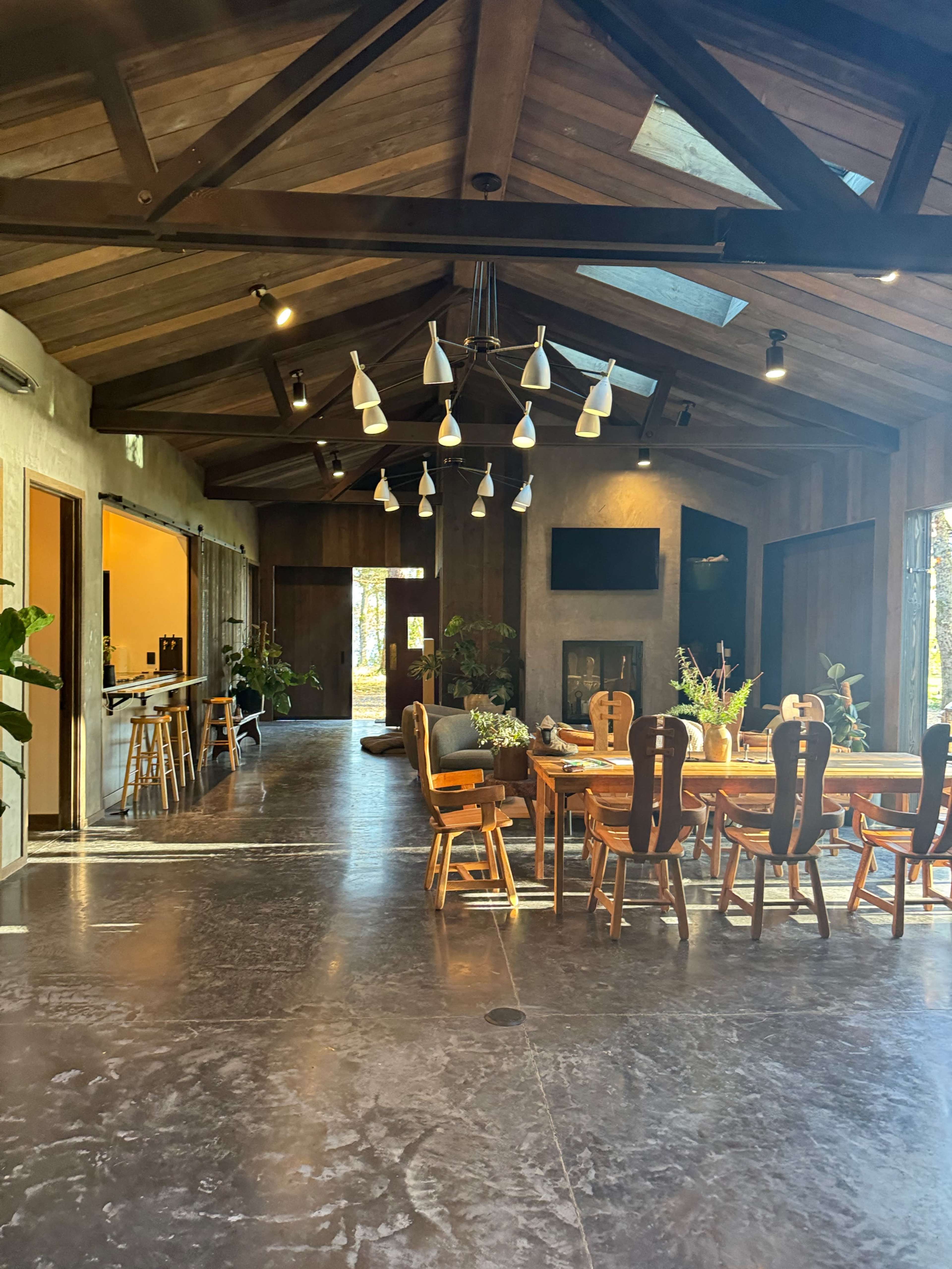 The image shows a spacious, modern interior with a large dining table surrounded by chairs, high wooden beams on the ceiling, and abundant natural light.