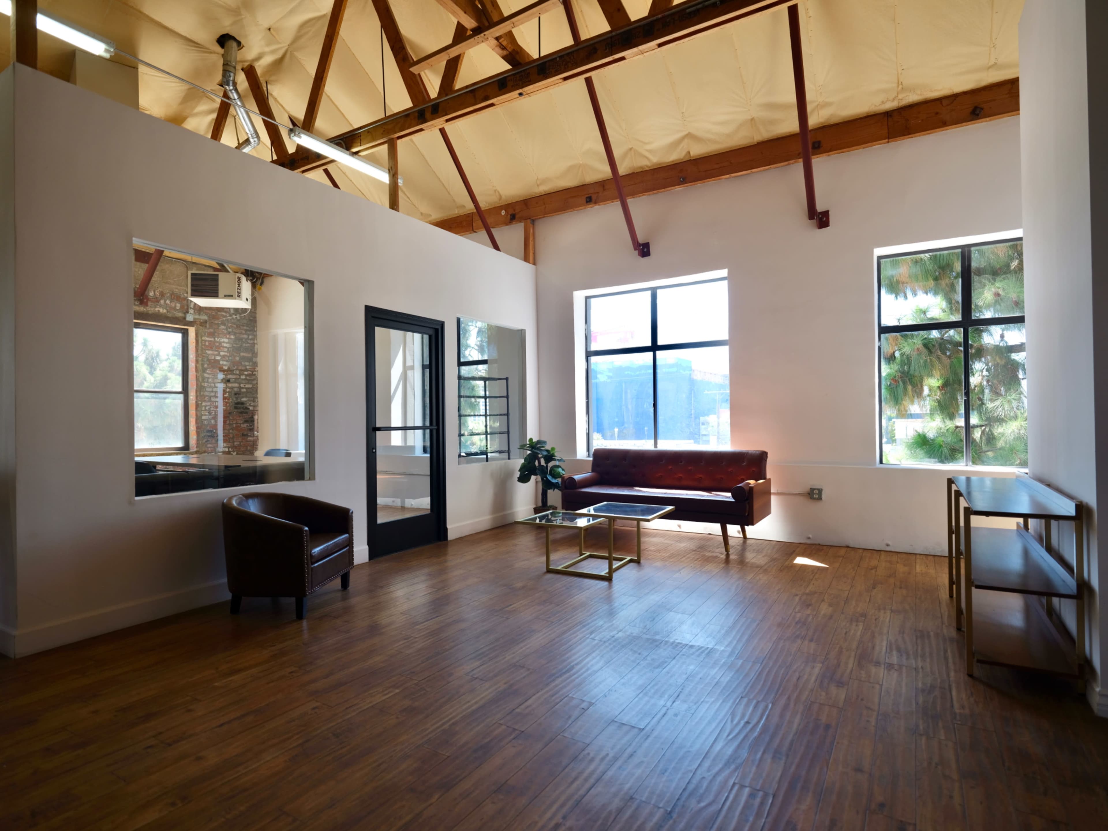 A spacious room features wooden flooring, large windows, a brown sofa, a glass coffee table, and a chair, with exposed beams on the ceiling.