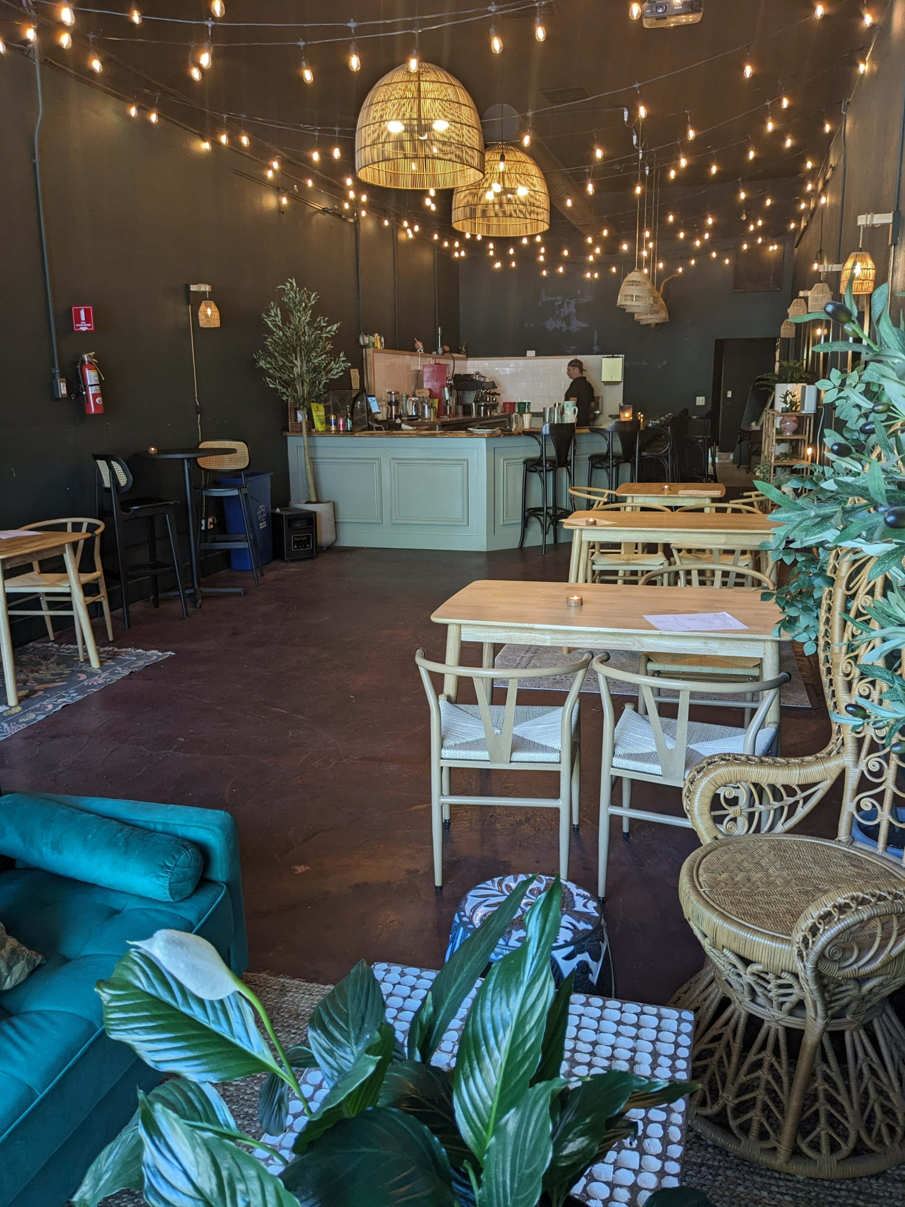 A spacious cafe features wooden tables and chairs, overhead light fixtures, and a counter with a barista in the background.
