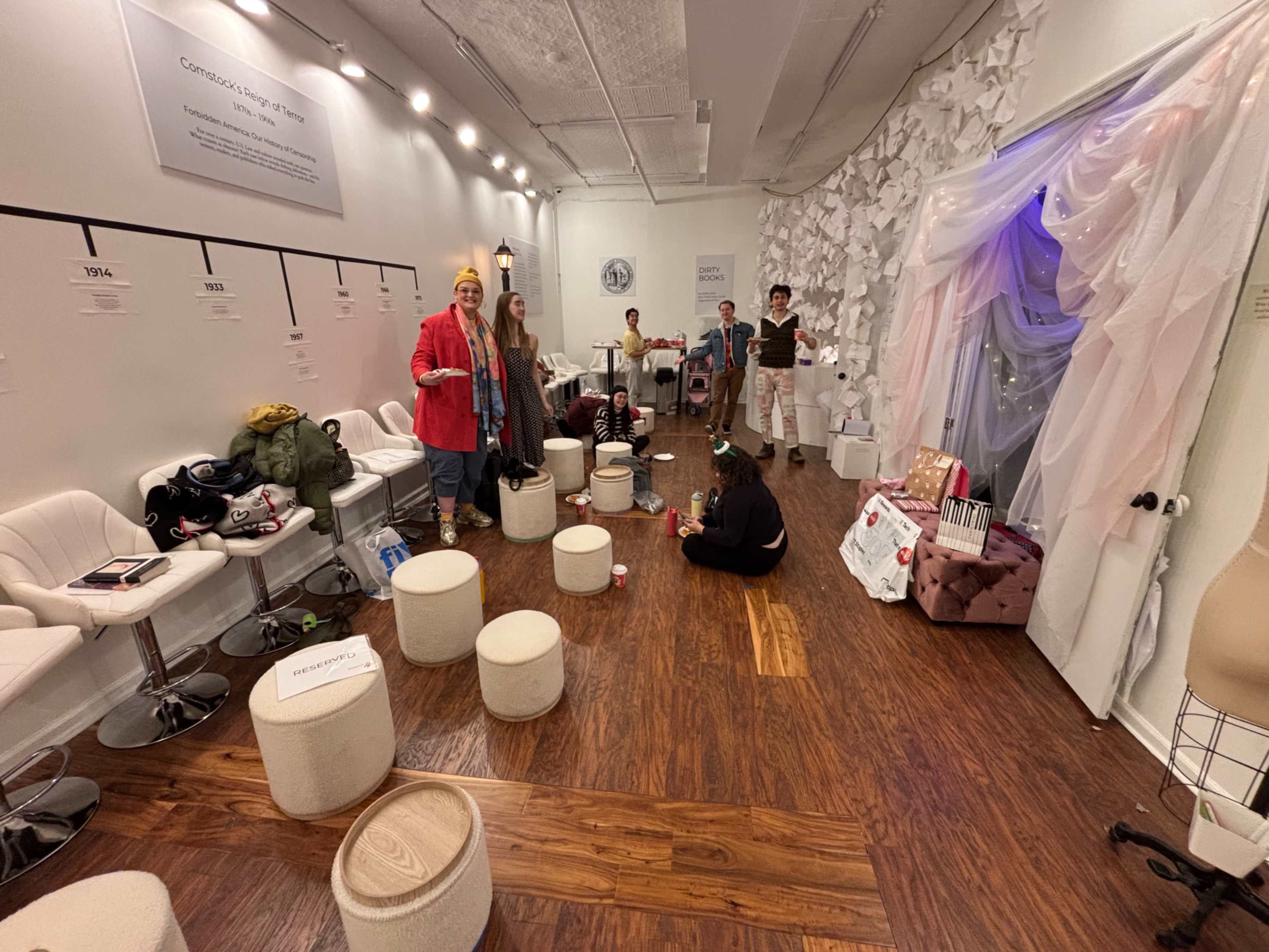 A group of people stands and sits in a brightly lit room with white walls, stylish seating, and decorative fabric draping.