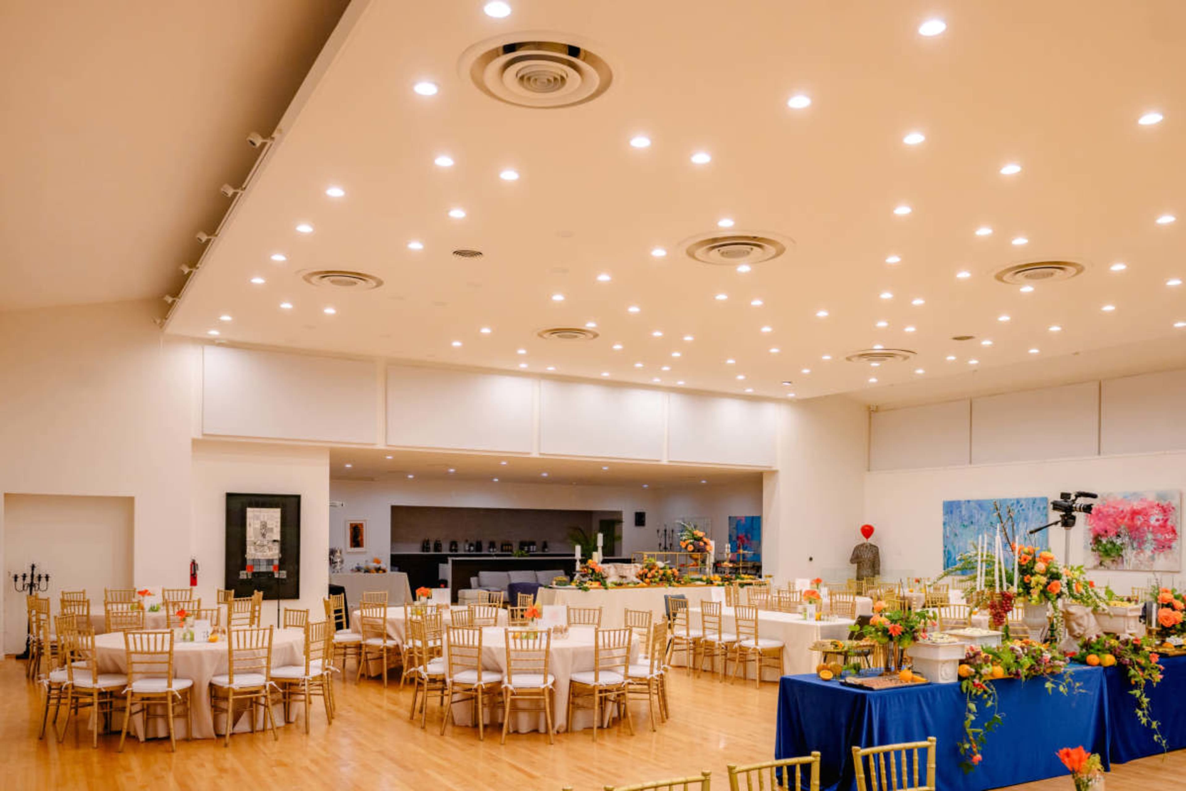 A spacious event hall features round tables with chairs arranged neatly, adorned with floral centerpieces under bright ceiling lights.