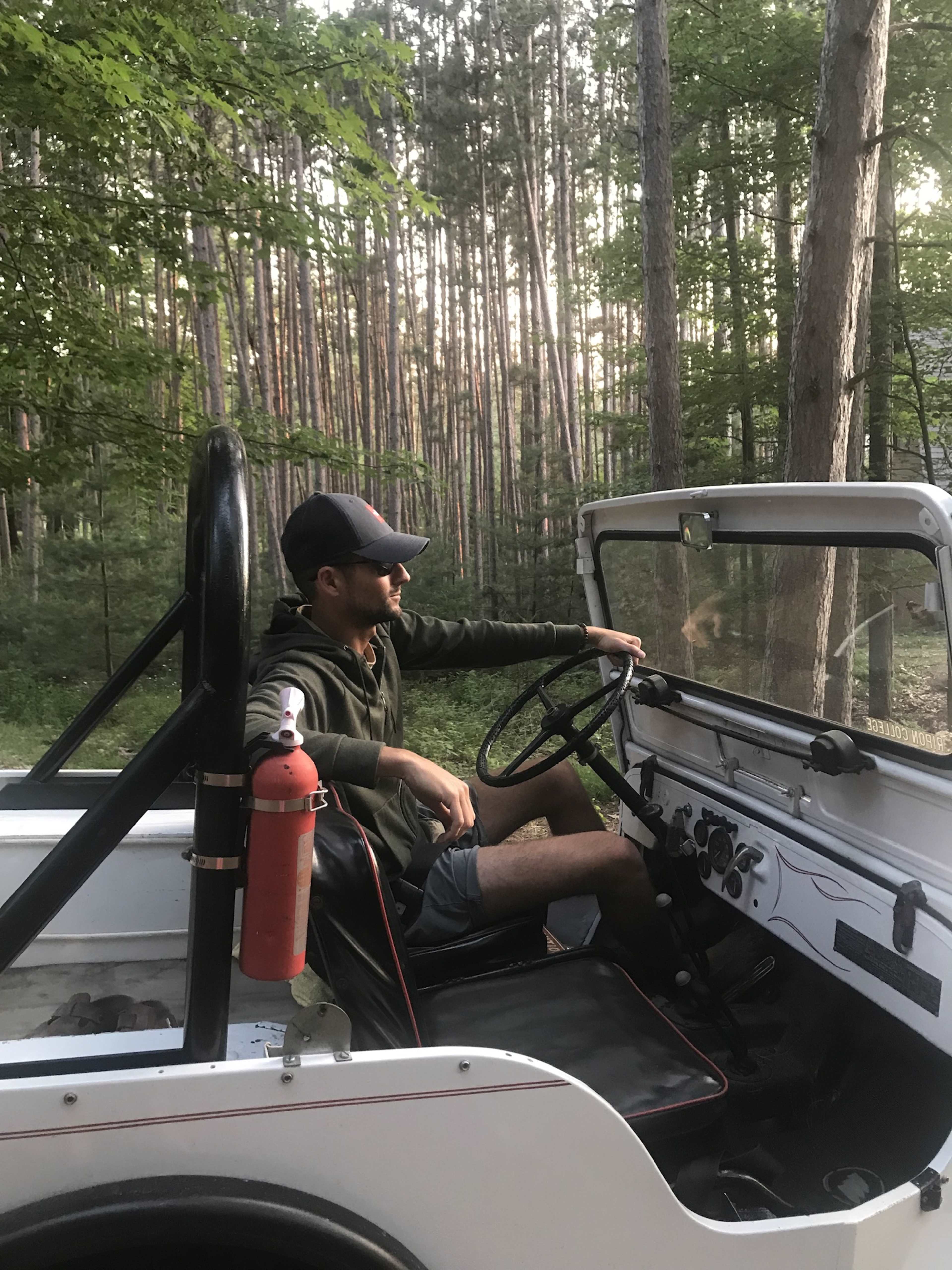 A man sits in the driver's seat of a jeep parked in a forest, surrounded by tall trees.