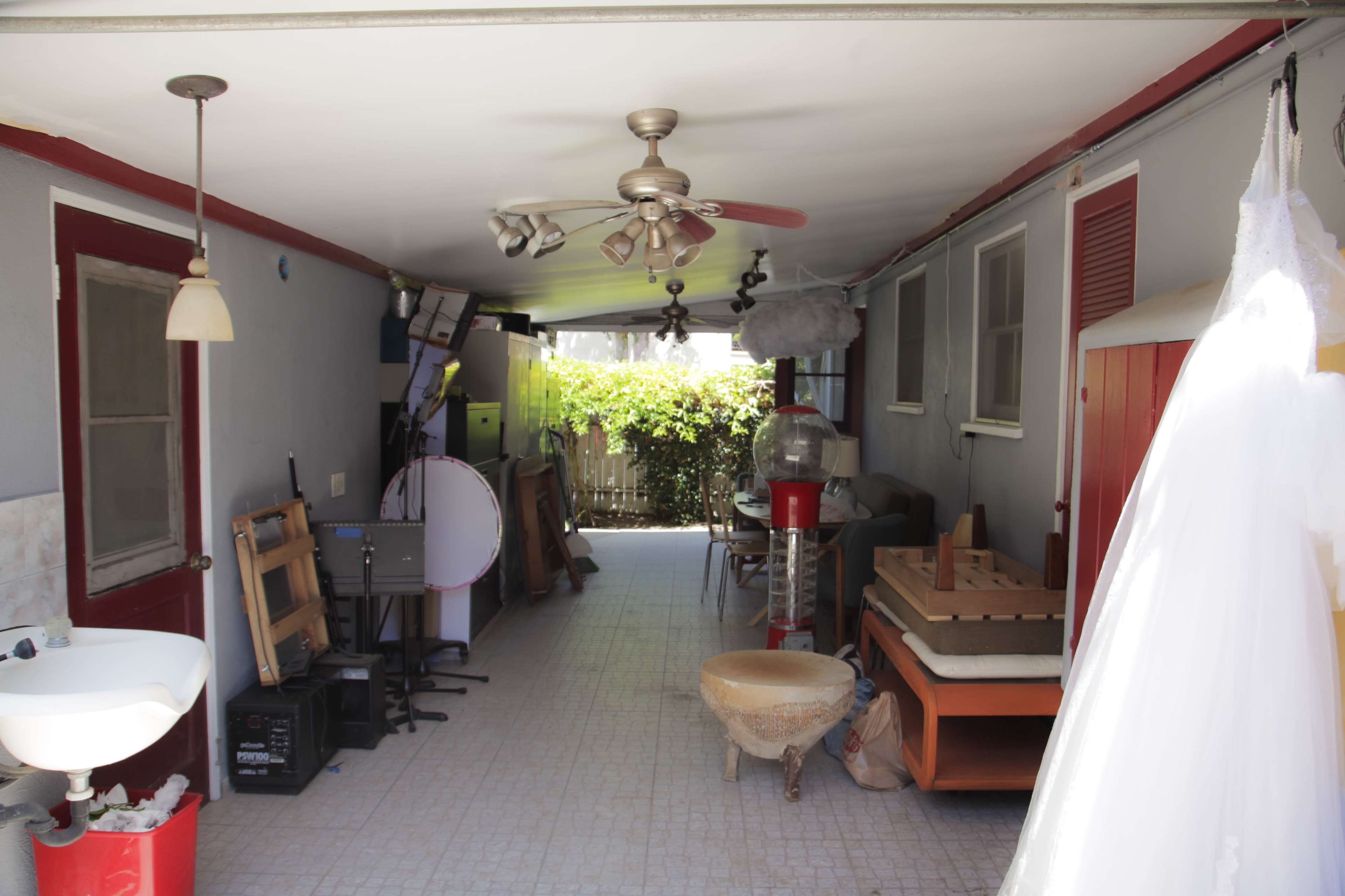 A narrow garage-like space lined with various items, including furniture, a fan, and a sink, with greenery visible outside.