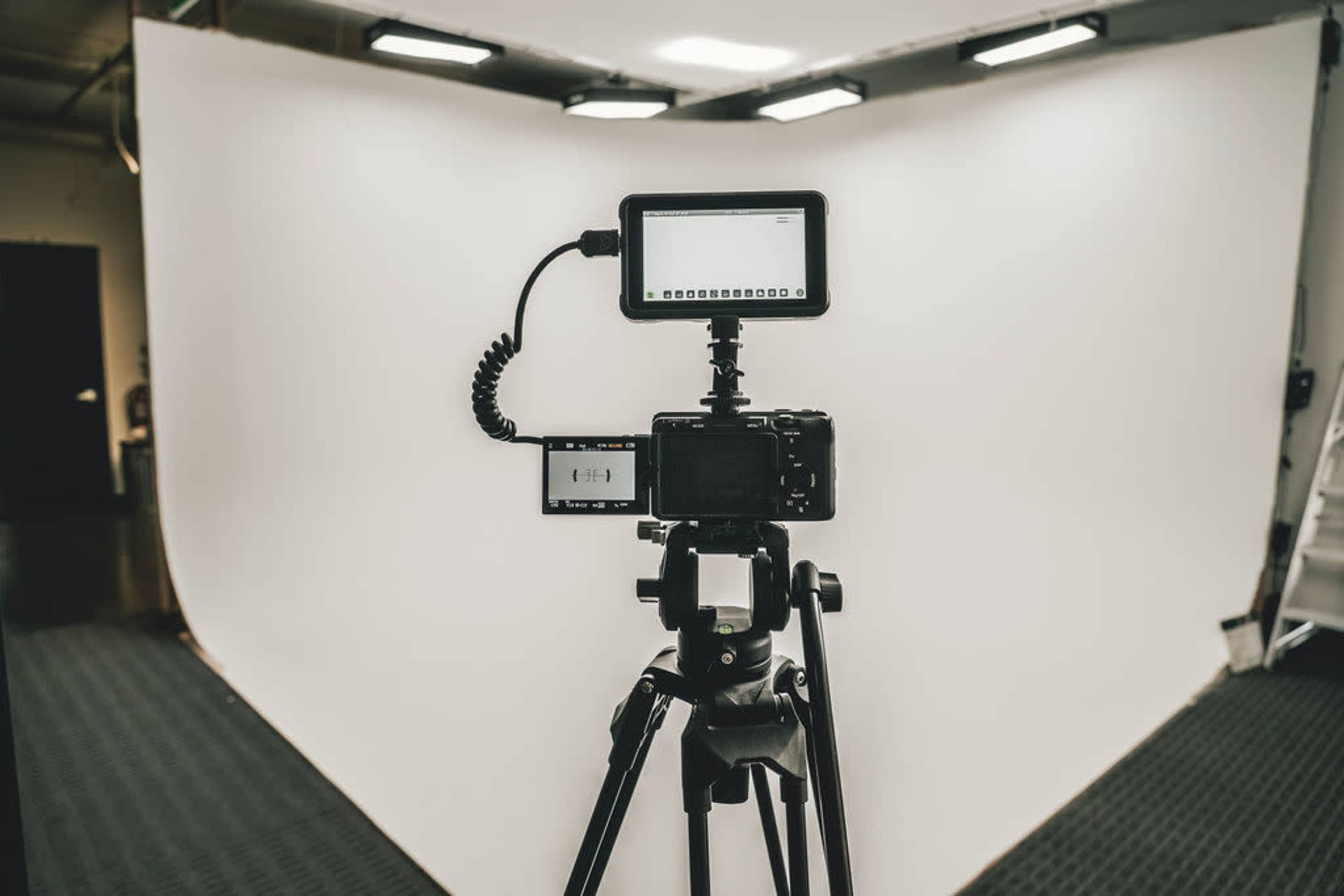 A camera mounted on a tripod is positioned in front of a blank white backdrop, with an external monitor connected to it.