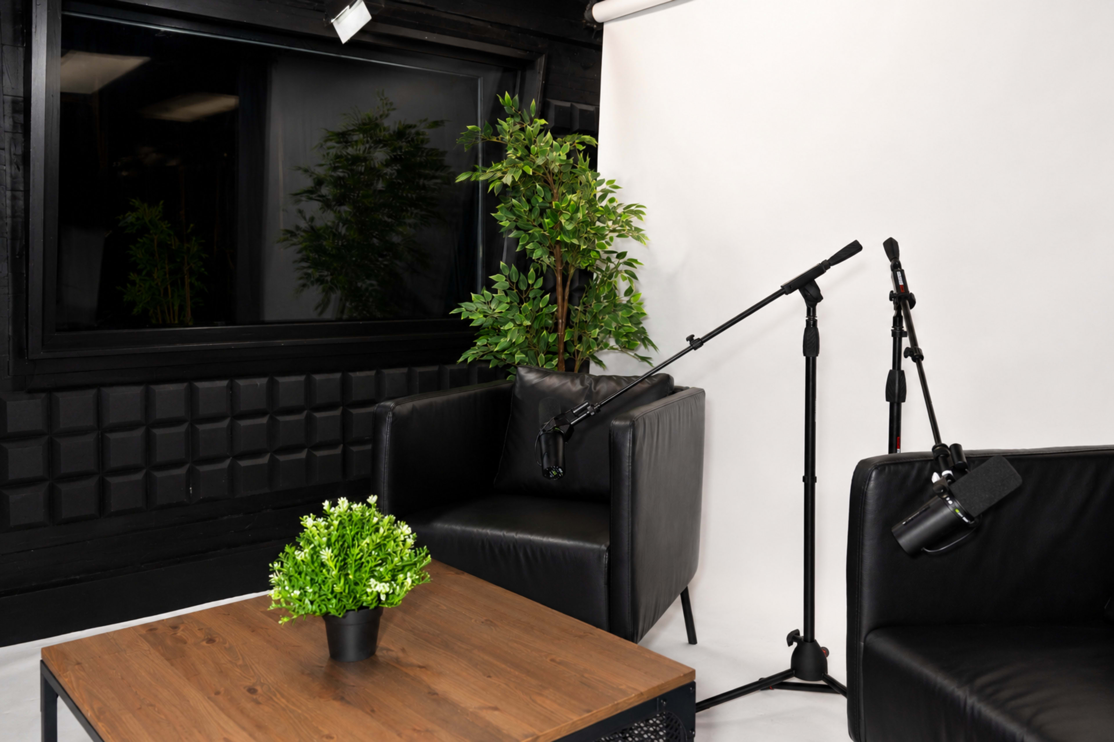 The image shows a small studio setup with two black chairs, a wooden table, and microphones positioned for recording.