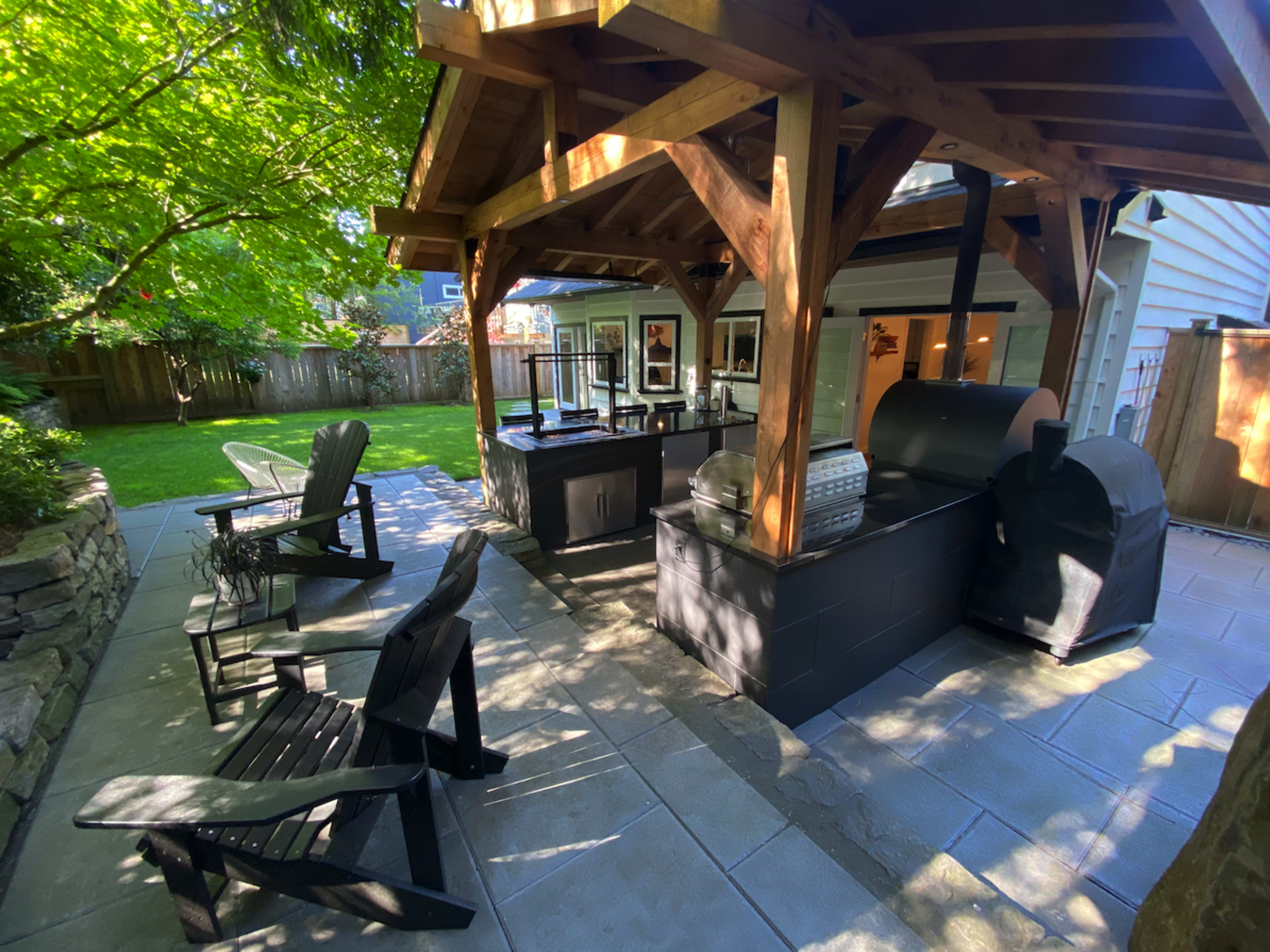 The image shows an outdoor kitchen area with a covered grill and seating space surrounded by greenery.