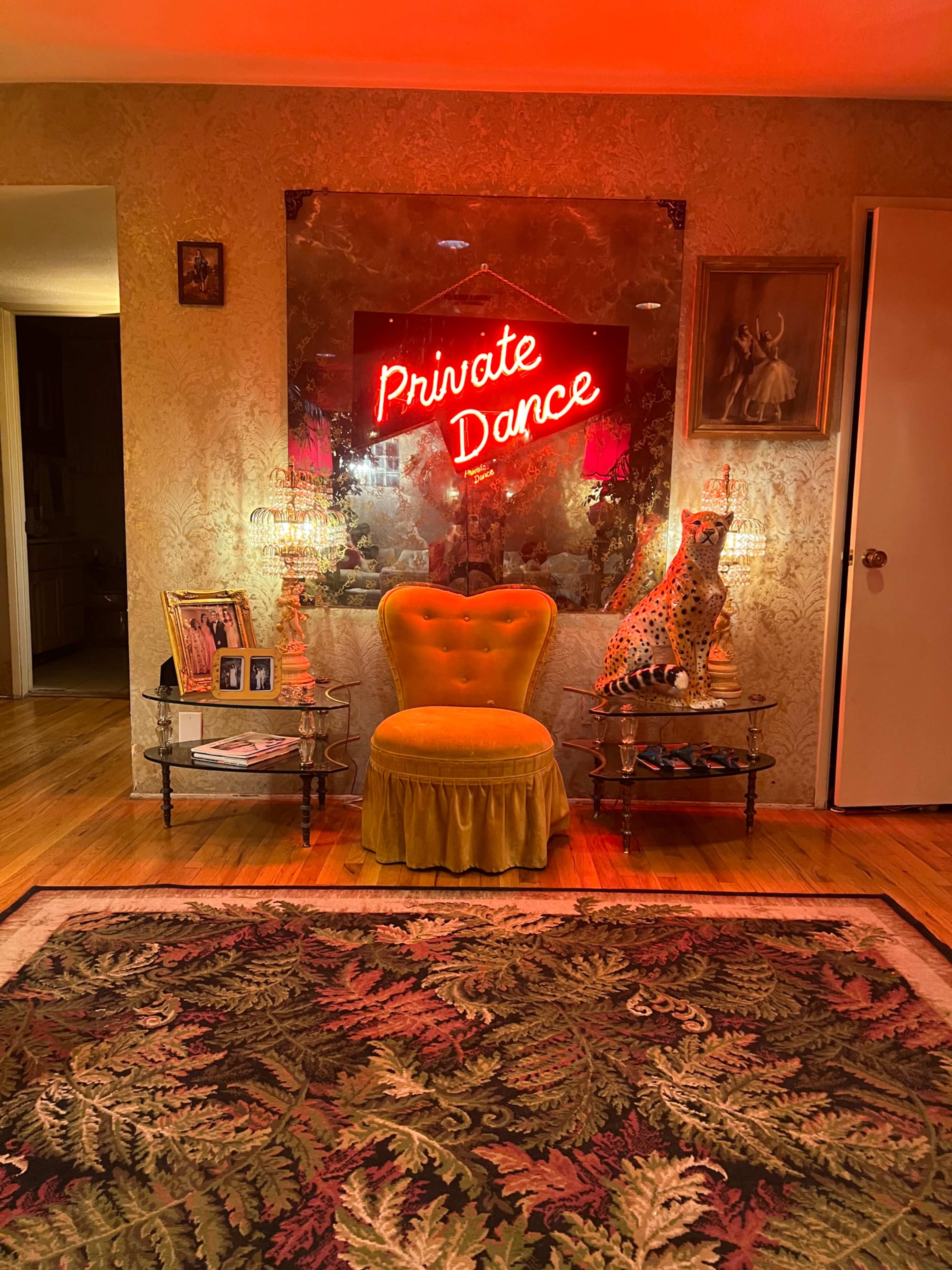 A yellow chair sits in front of a neon sign reading "Private Dance," surrounded by a decorative rug and various decor elements in a warm-lit room.