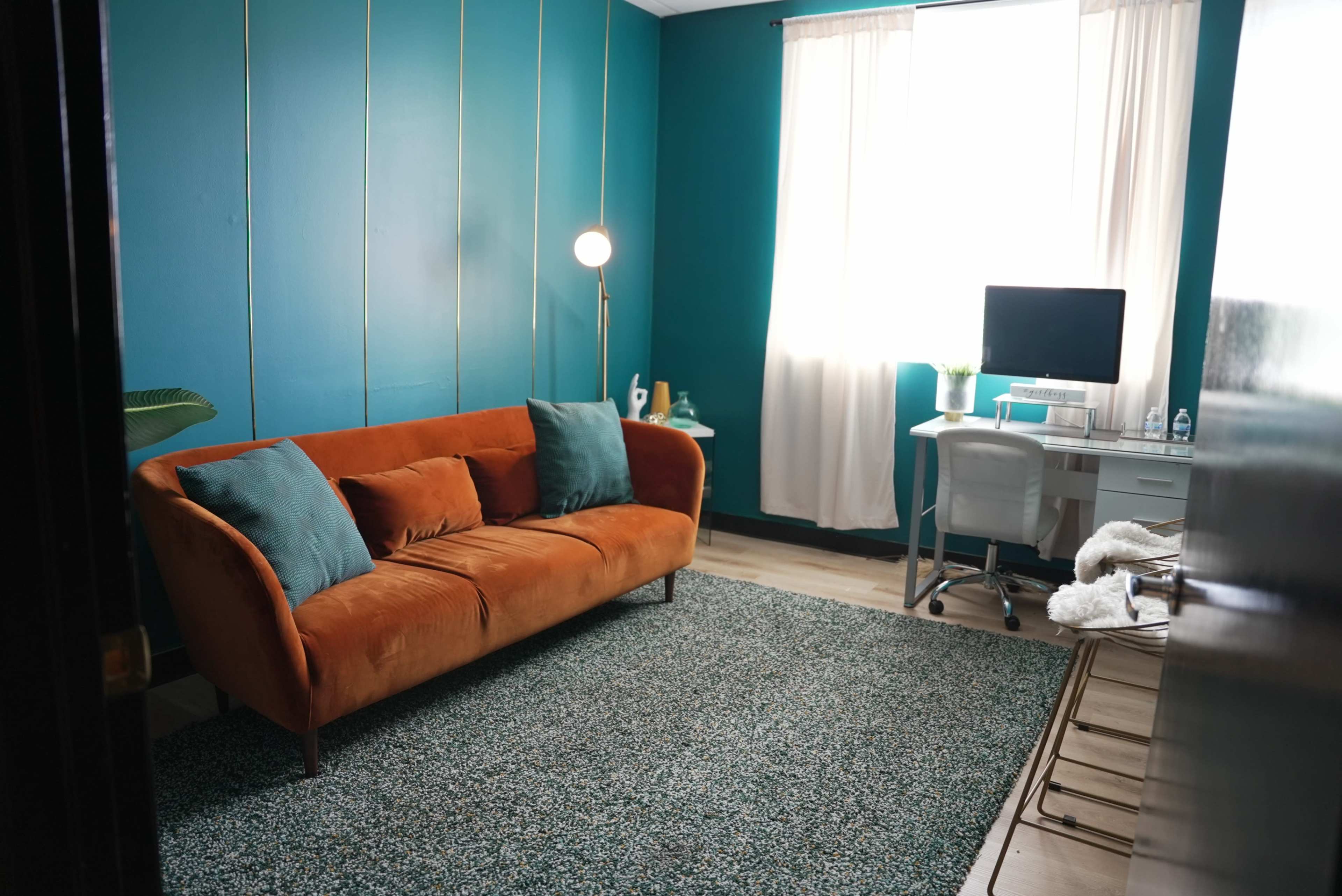 The image shows a cozy room featuring an orange velvet sofa, a green area rug, a desk with a computer, and large windows with white curtains.