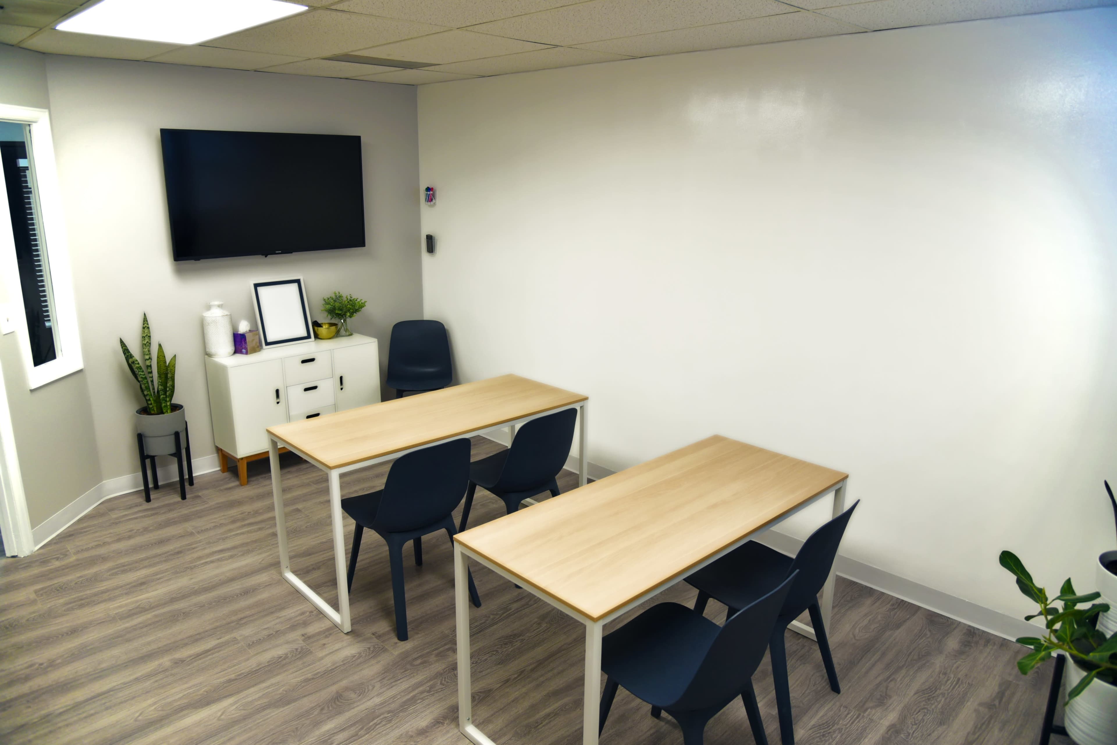 A meeting room with two wooden tables, four chairs, a wall-mounted TV, and a whiteboard.