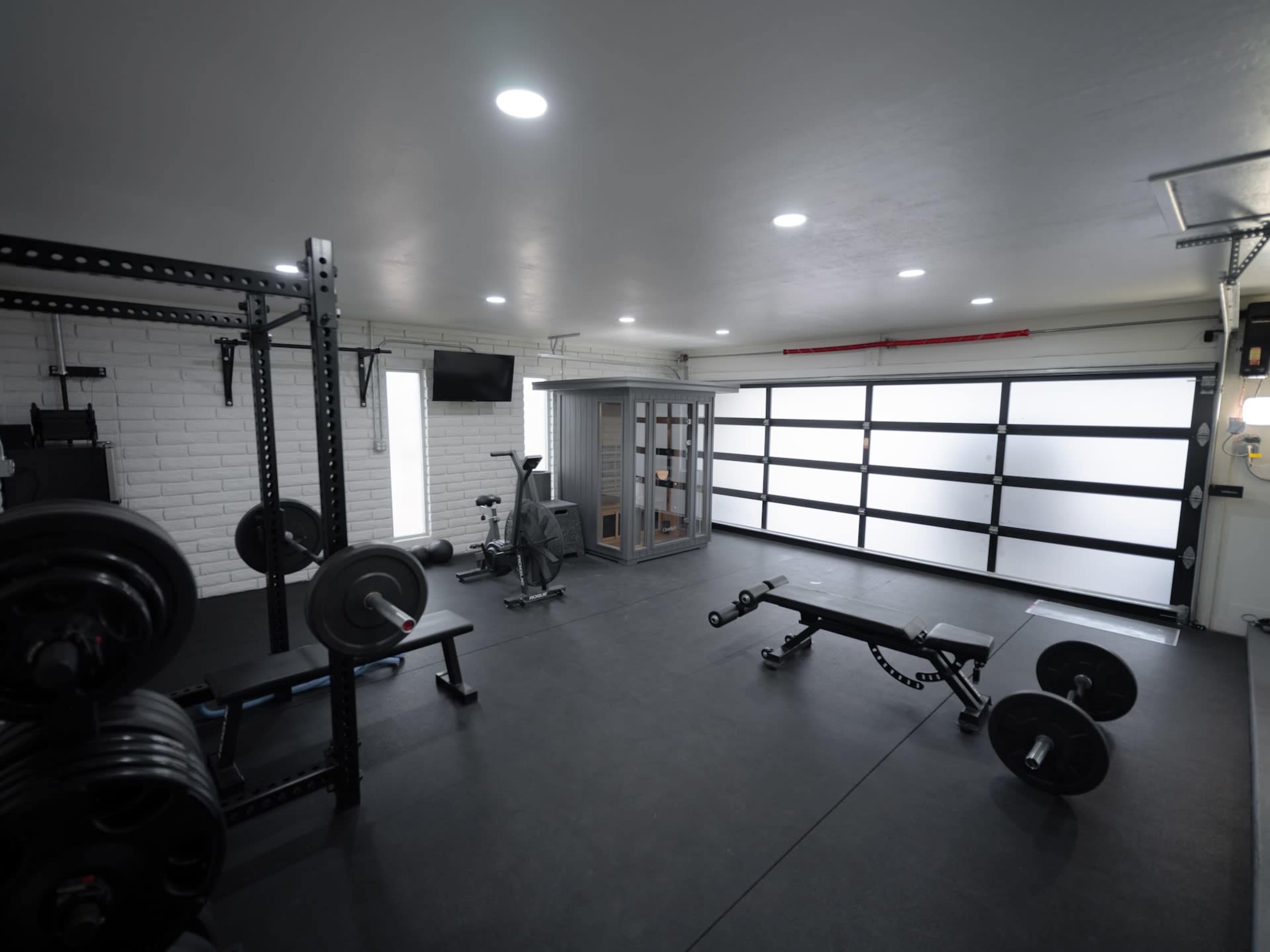 The image shows a spacious home gym with weightlifting equipment, a rowing machine, and a glass-enclosed sauna.