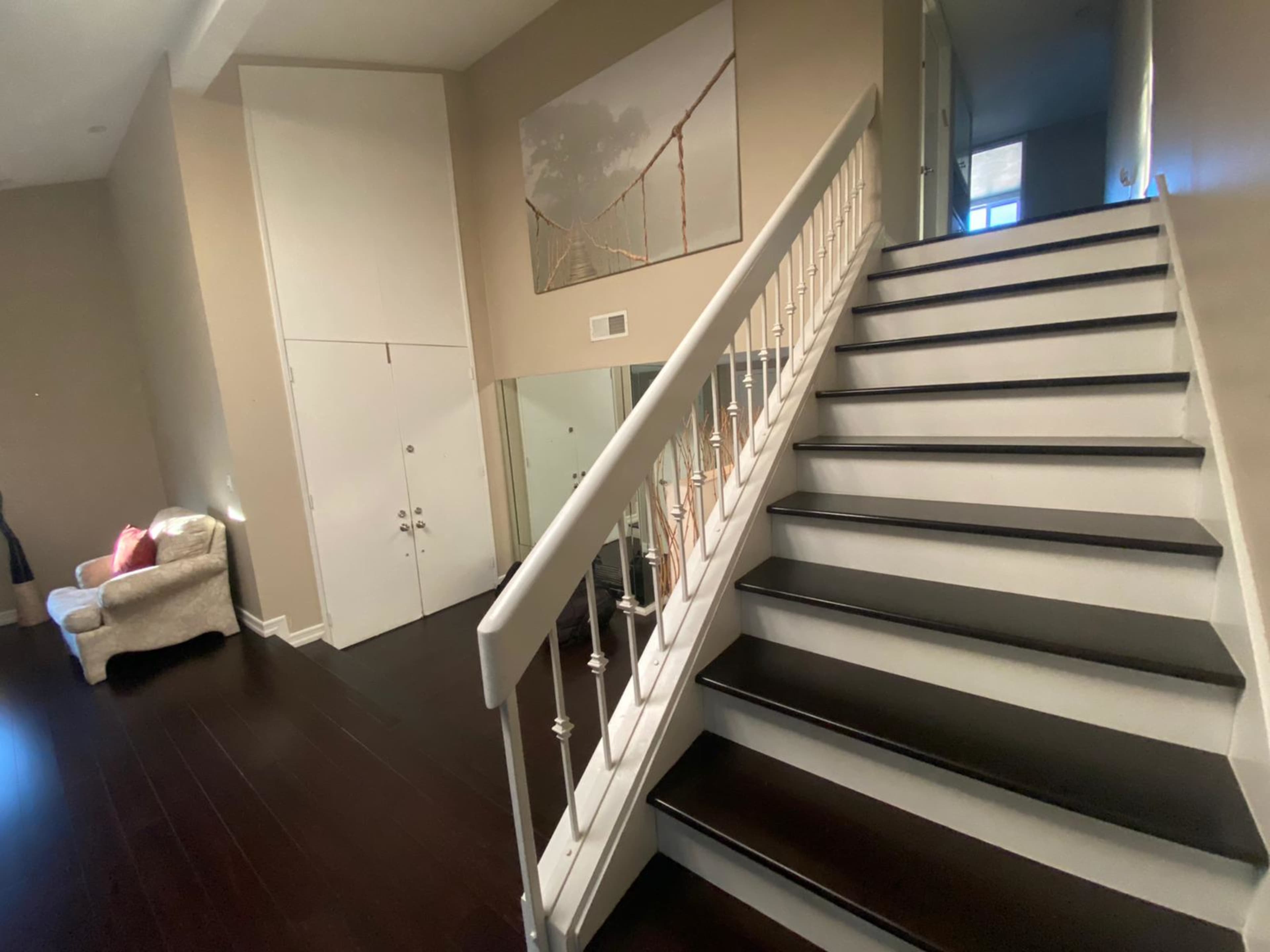 A staircase with white railings ascends in a well-lit foyer that features a large wall art piece and a beige seating area.