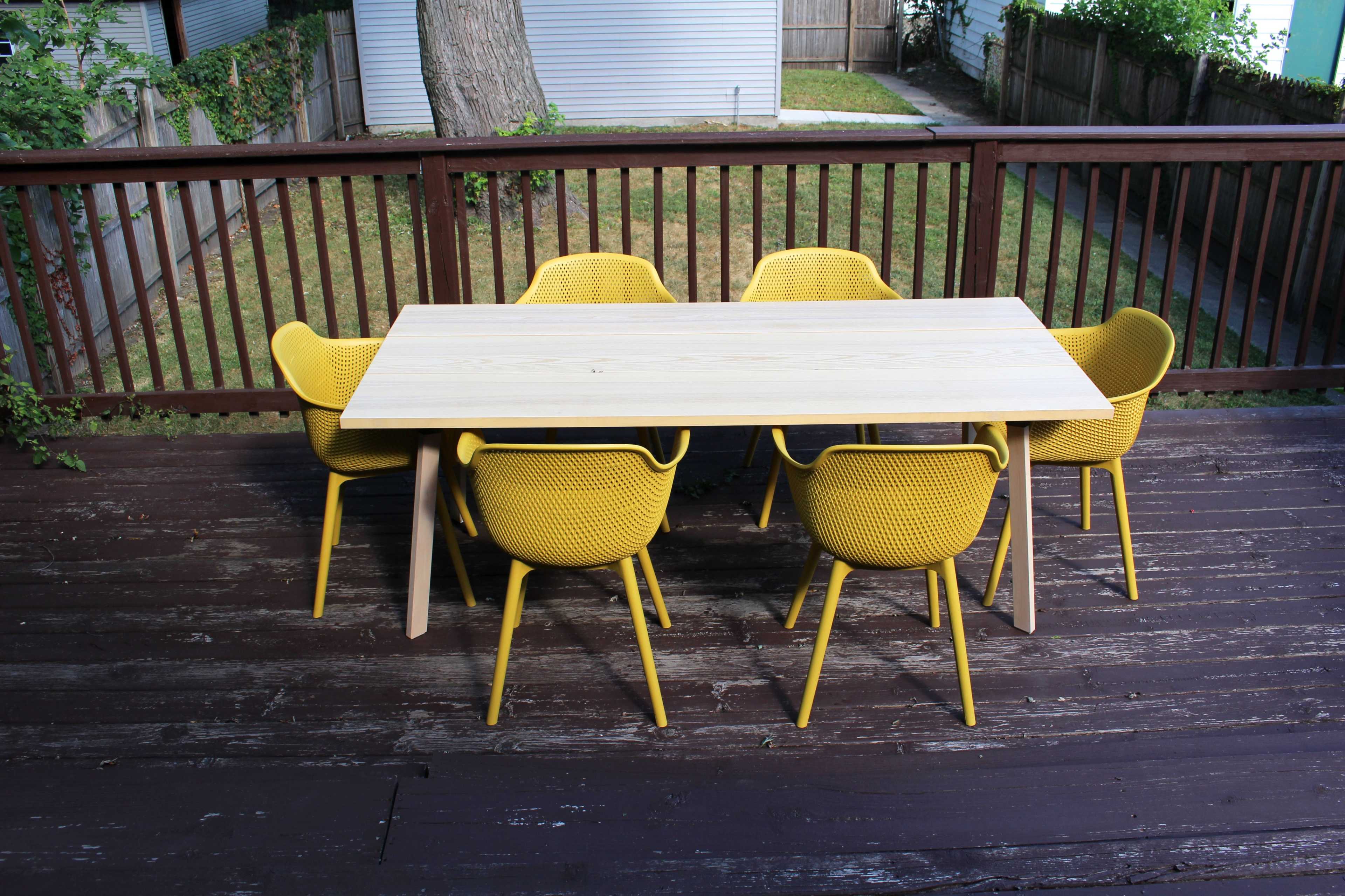 A wooden table surrounded by six yellow chairs is set on a deck with a view of a yard and fence.