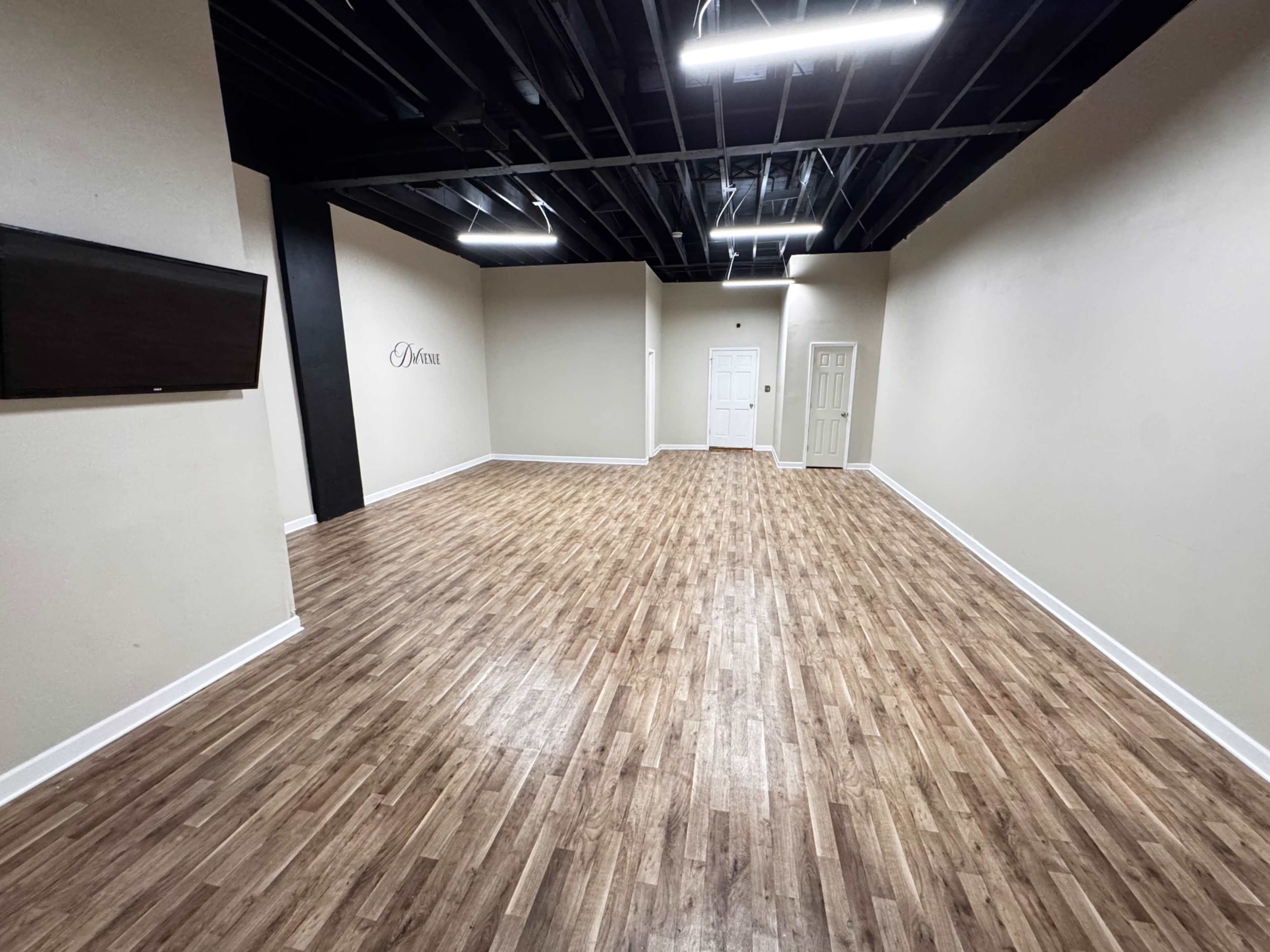 The image shows a spacious, empty room with wooden flooring, light-colored walls, and a door at the far end.