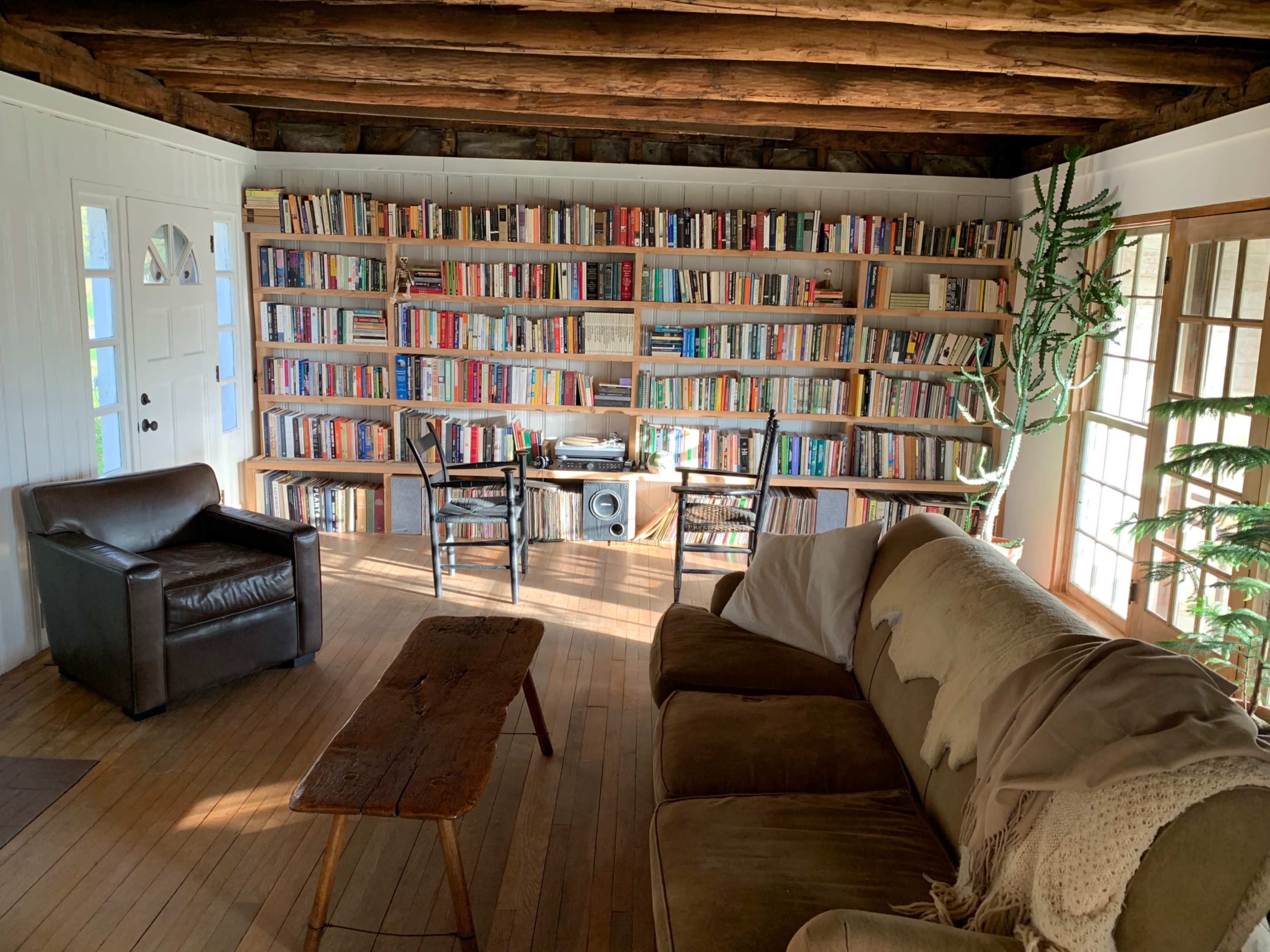 A cozy living space features a large bookshelf filled with books, a wooden coffee table, two chairs, and a sectional sofa with a throw blanket, illuminated by natural light.