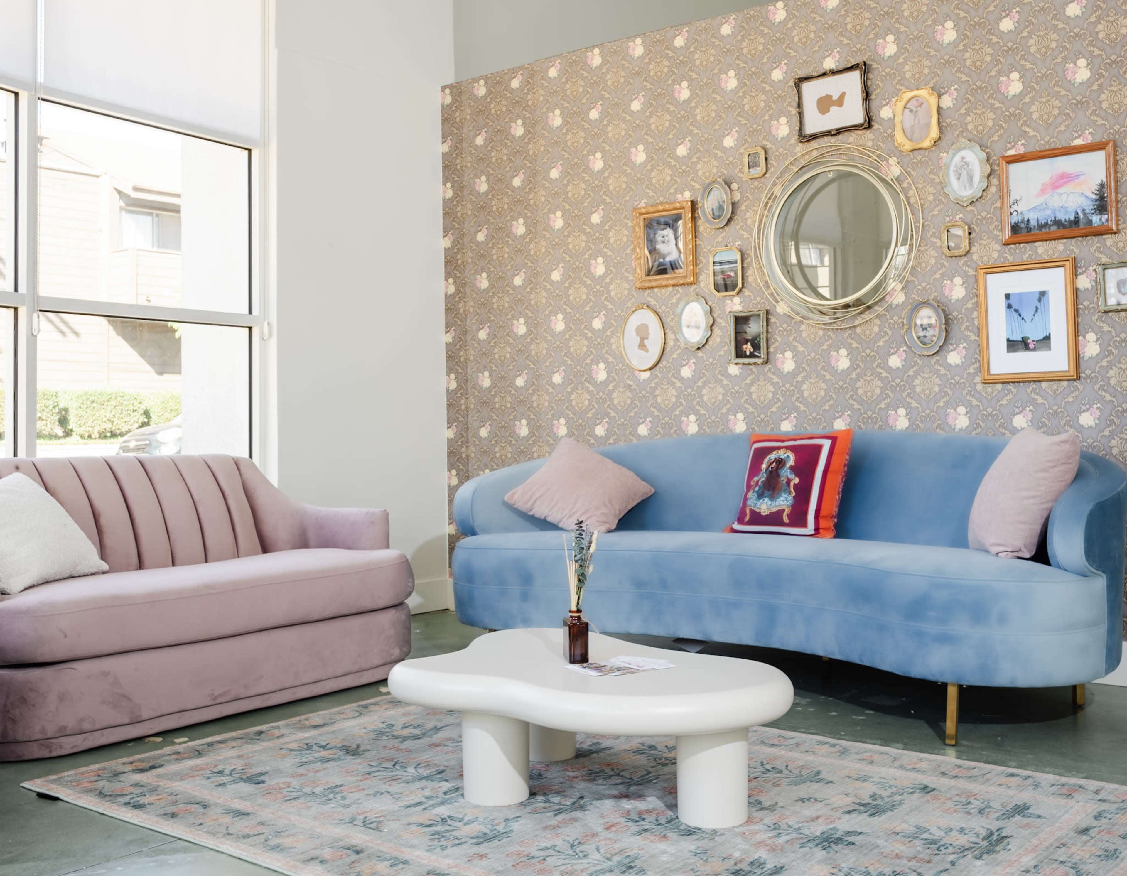 A modern living room with a pink sofa and a blue curved couch, accompanied by a round mirror and framed pictures on a patterned wallpaper backdrop.