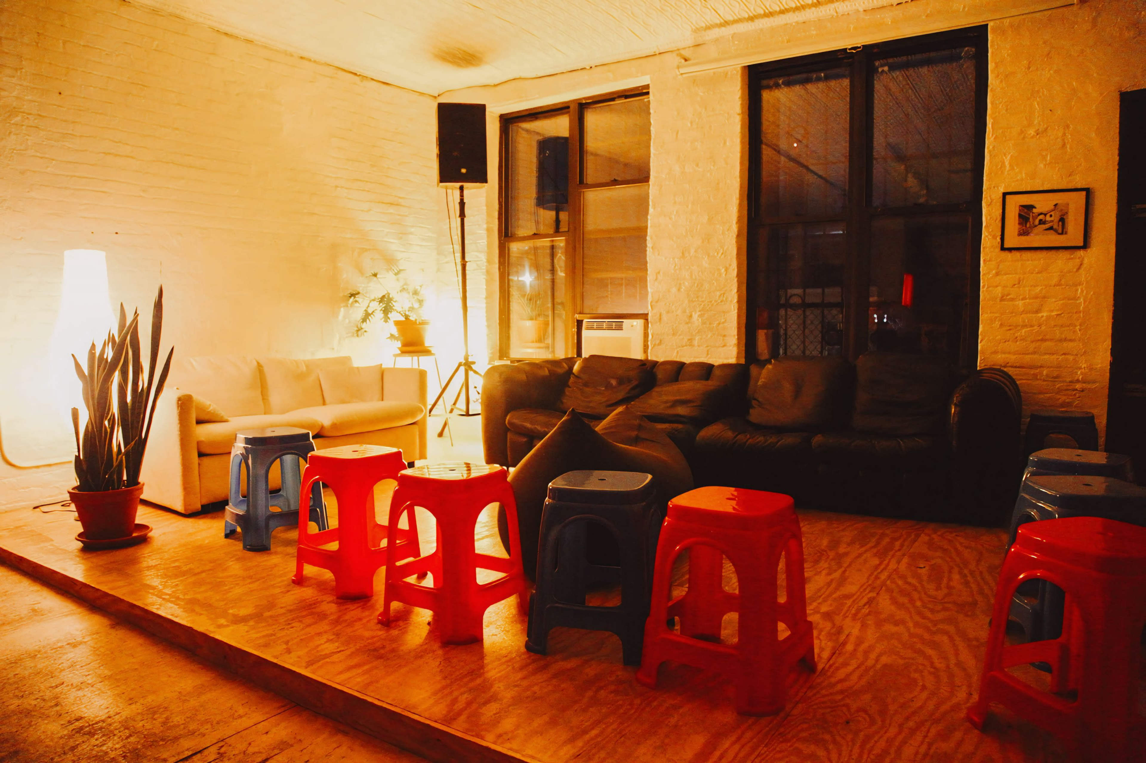 The image shows a cozy indoor space with a mix of seating options, including a white couch, a black leather sofa, and colorful plastic stools arranged in a line on a wooden platform.