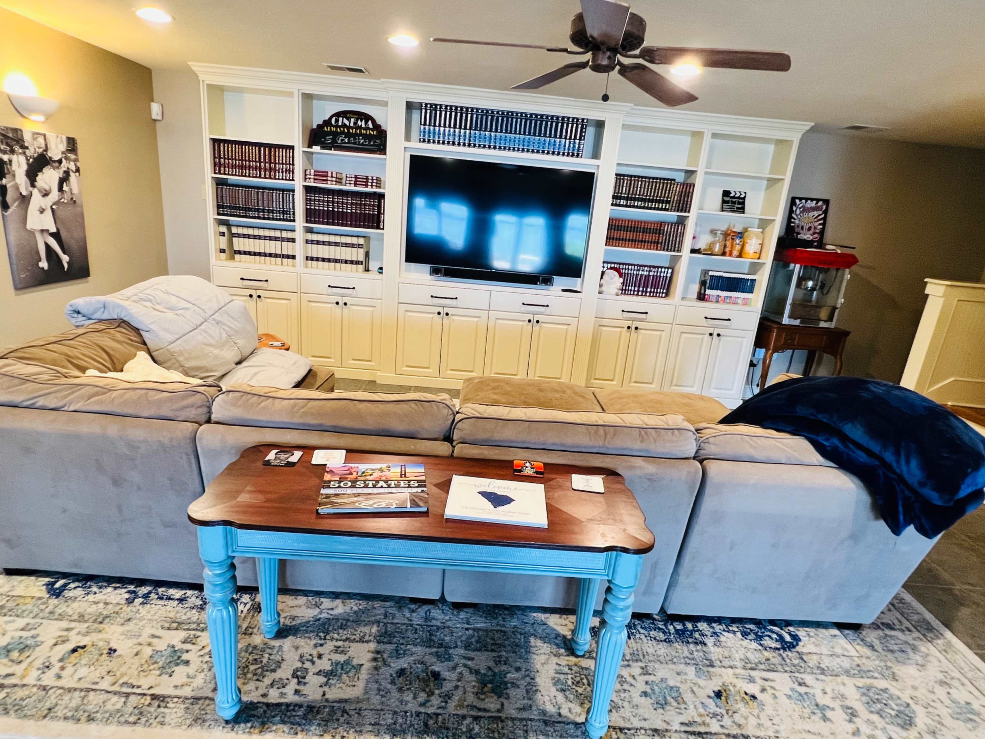 A living room features a large sectional sofa facing a white entertainment unit filled with books, a flat-screen TV, and a coffee table in front.
