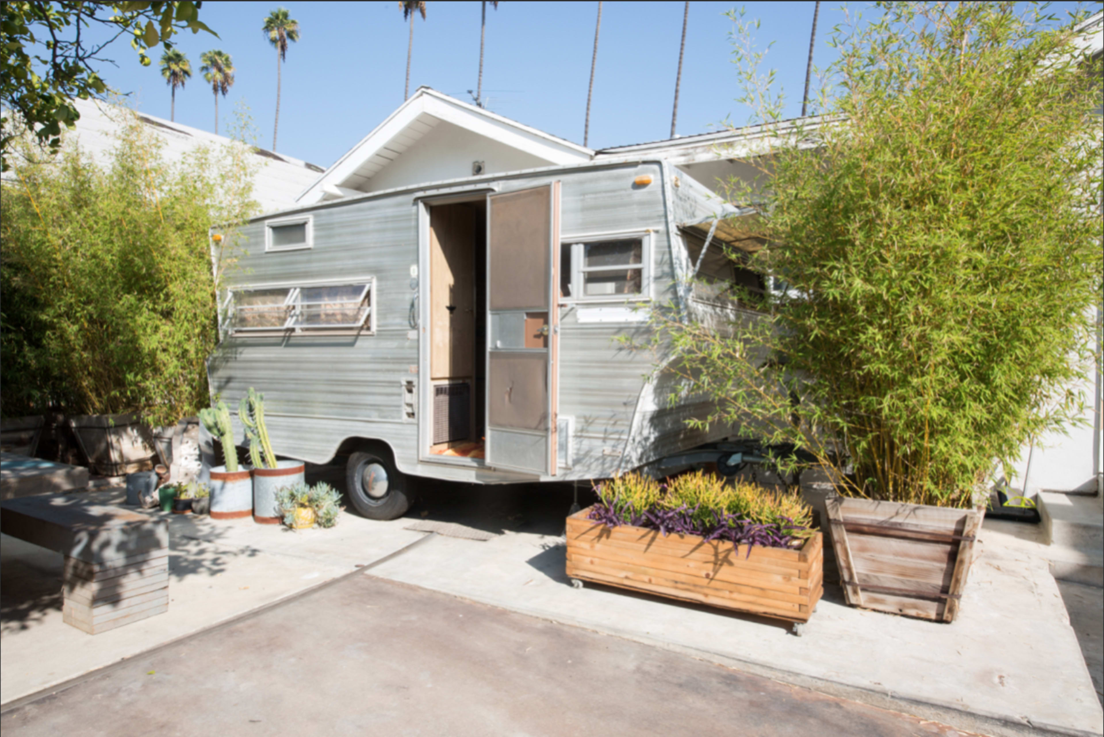 A vintage camper trailer is parked beside a modern house, surrounded by potted plants and bamboo.