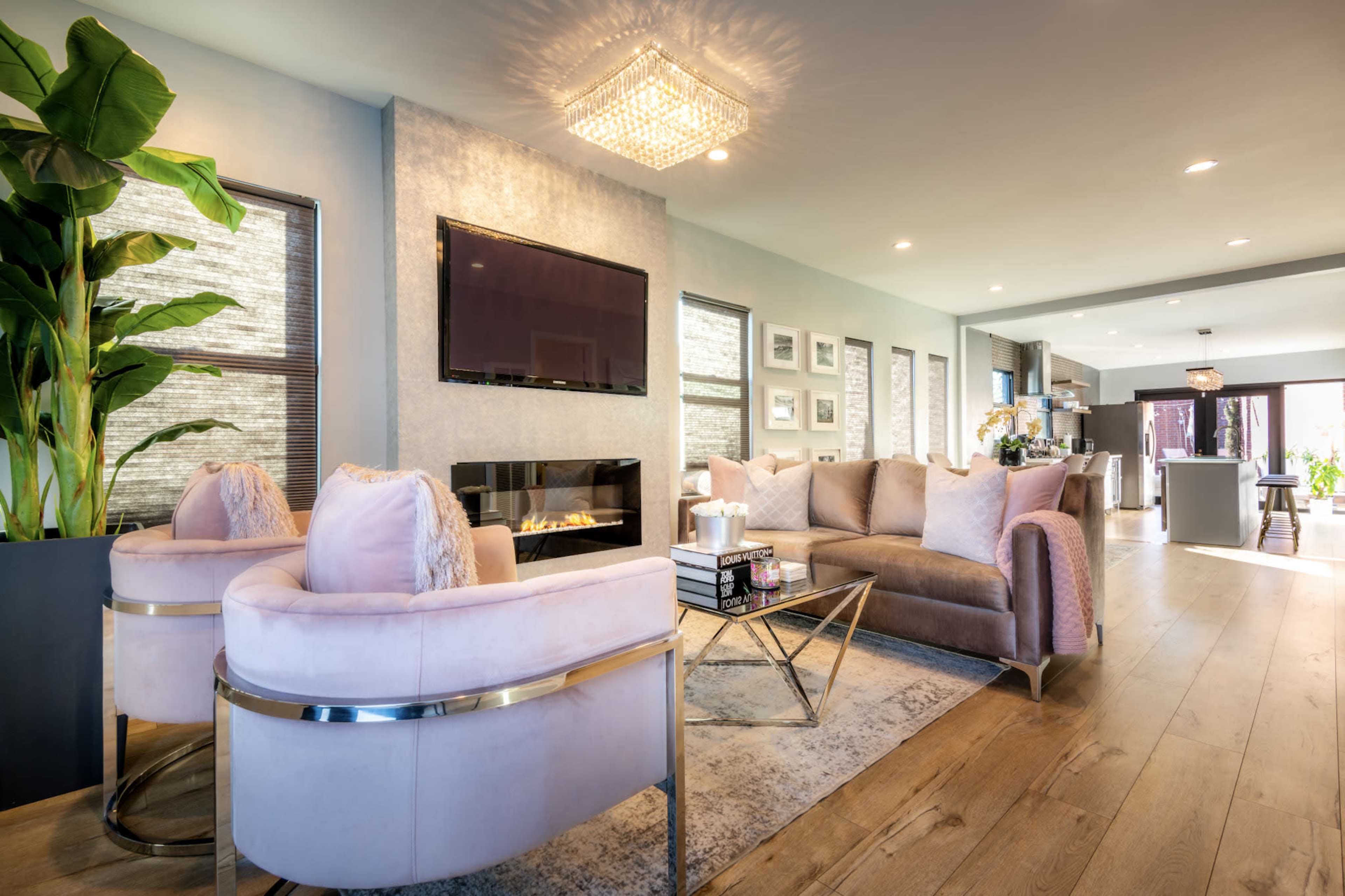 A modern living room featuring a light pink sectional sofa, two matching accent chairs, a flat-screen TV mounted on the wall, and a fireplace, all surrounded by large windows and decorative plants.