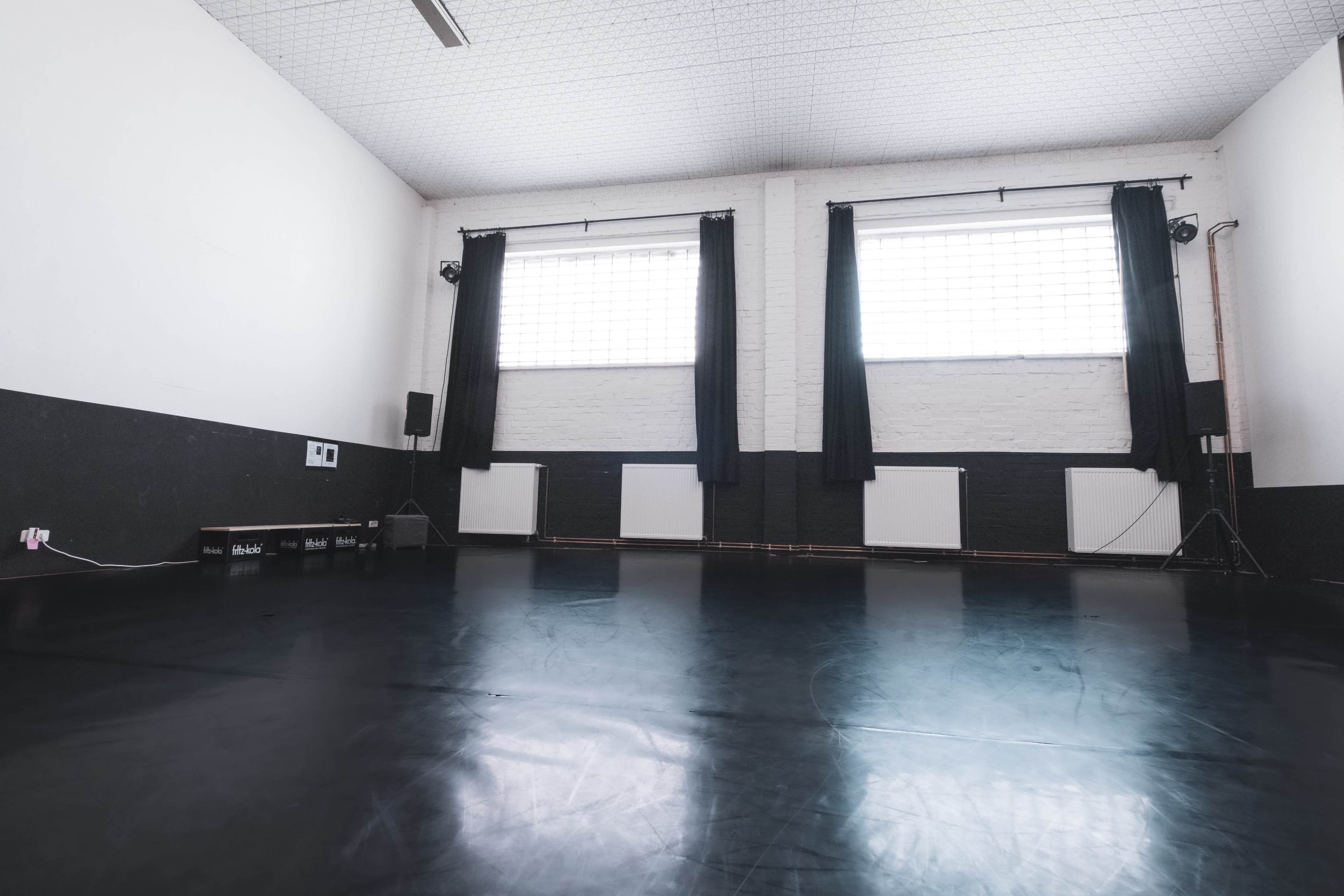 An empty dance studio with dark flooring and large windows covered by black curtains.