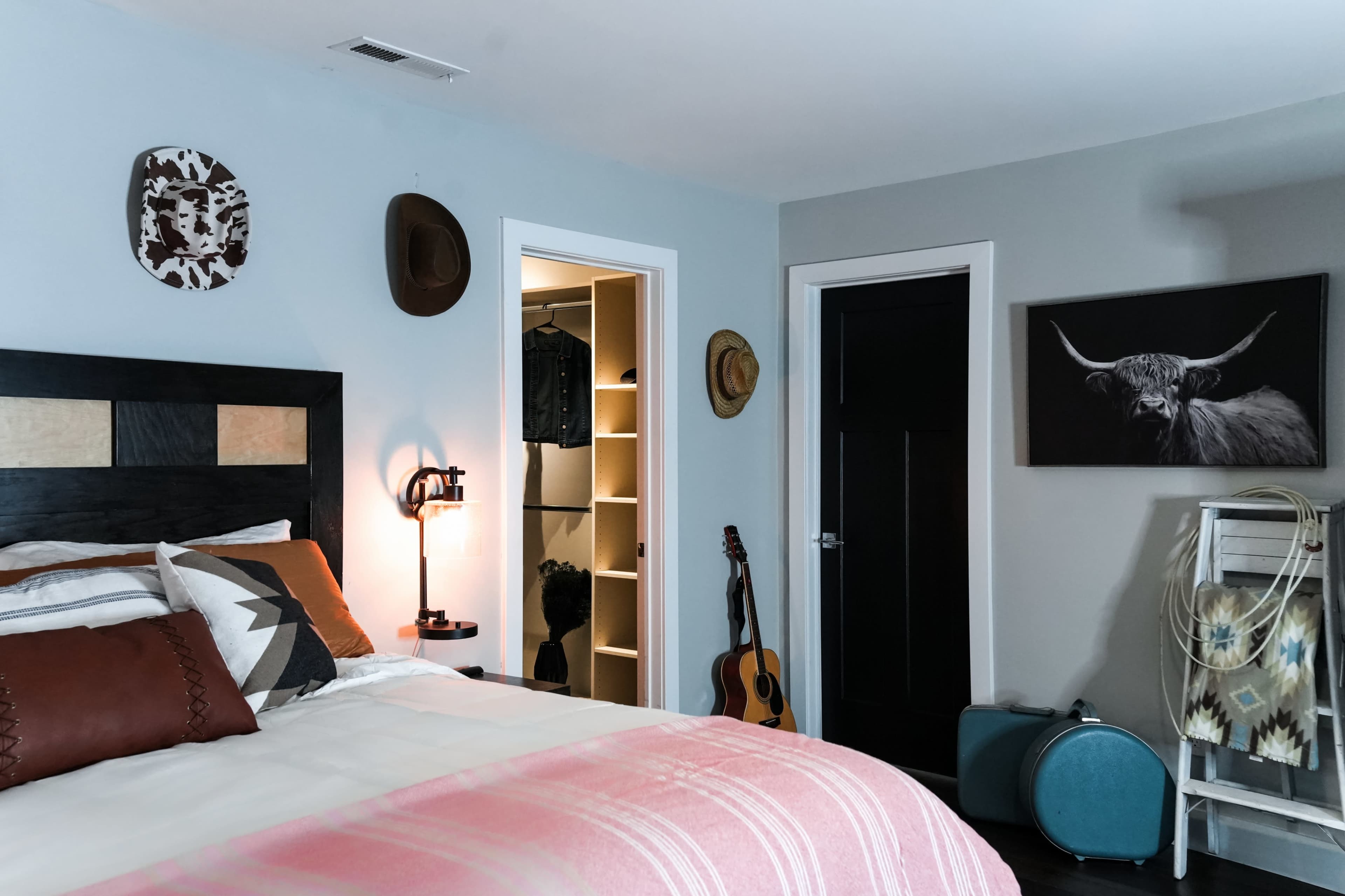 The image shows a modern bedroom featuring a bed with decorative pillows, an open closet revealing clothes, a guitar leaning against the wall, and artwork of a bull's head.