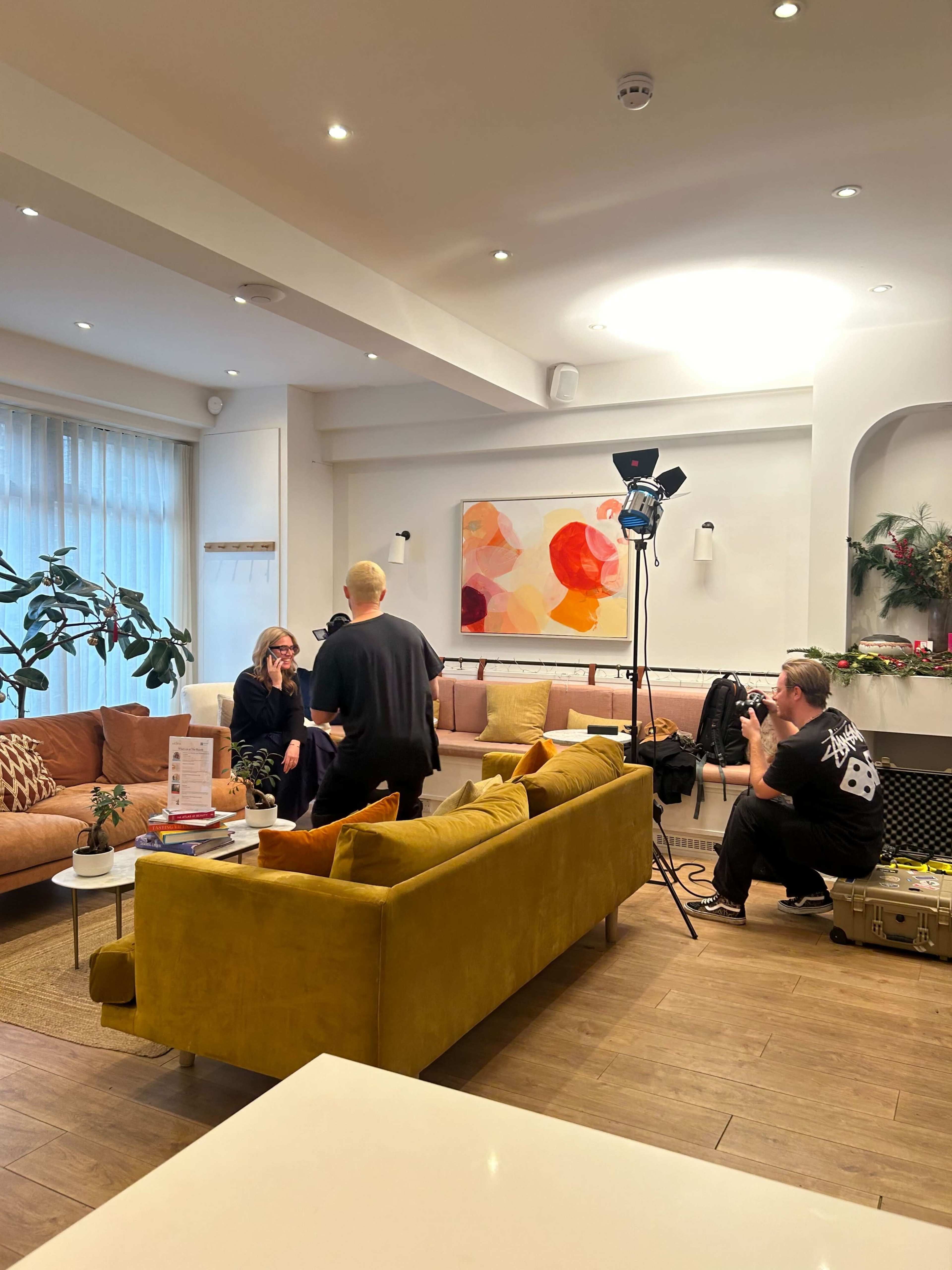 A person is sitting on a couch while another person prepares filming equipment in a well-lit living room.