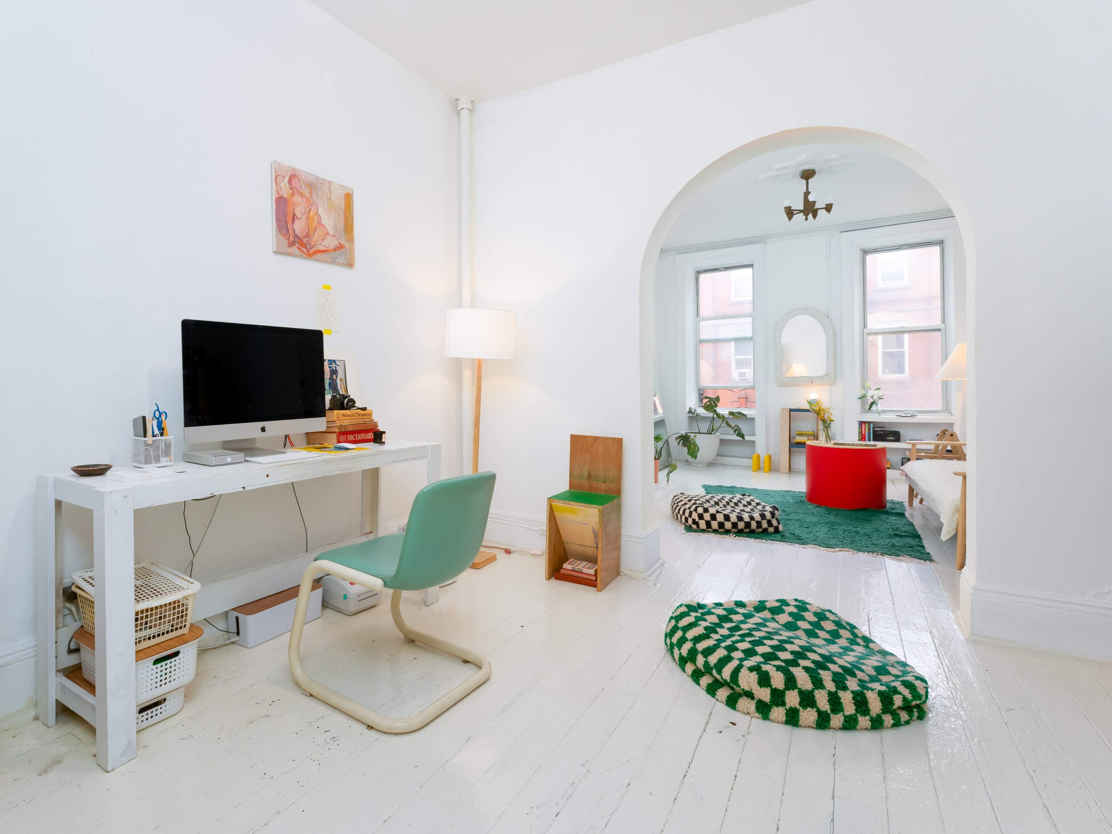 The image shows a bright, minimalist room with a white desk, a computer, a green chair, and a cozy seating area featuring a red and green rug.