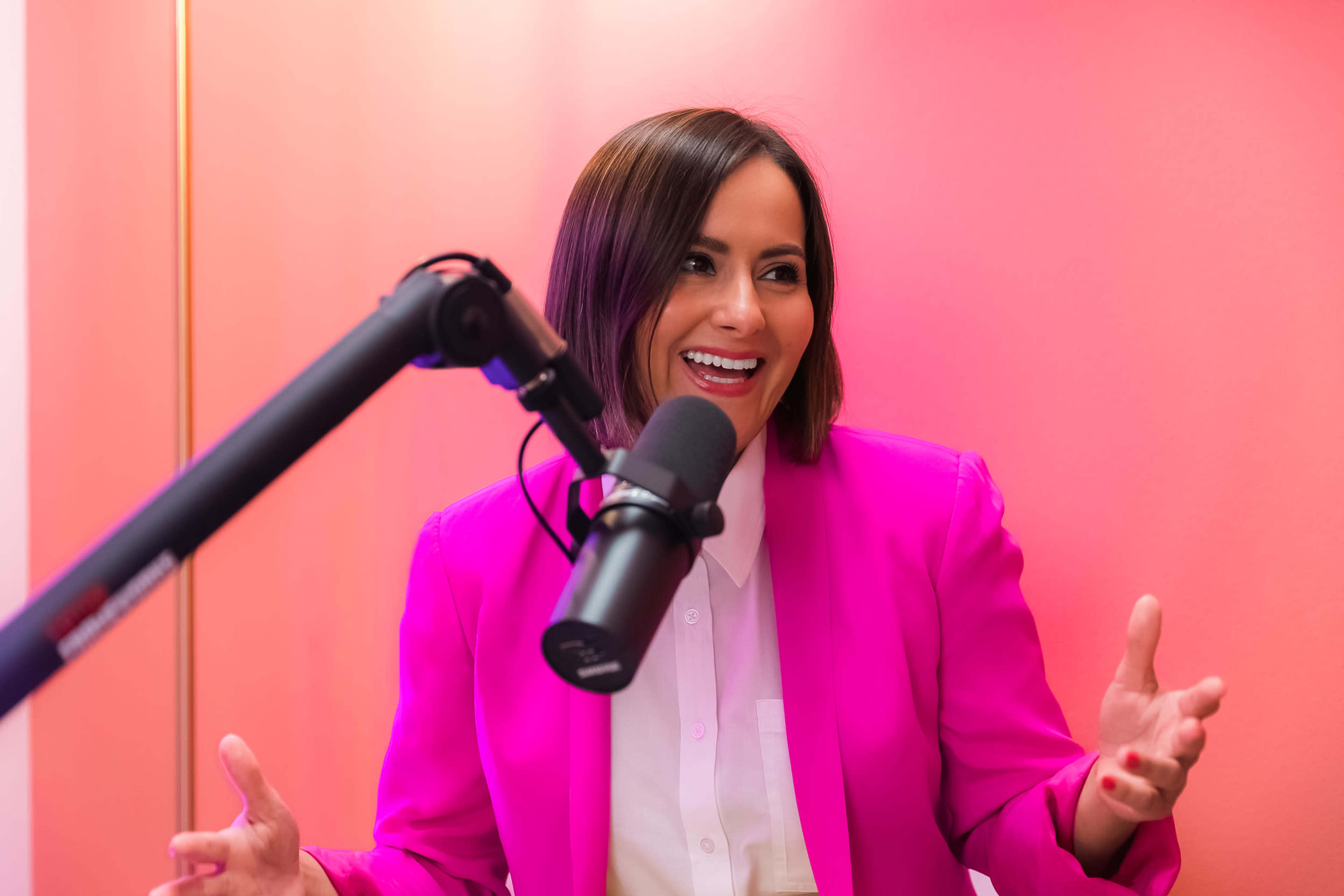 A woman in a bright pink blazer sits in front of a microphone, gesturing as she speaks against a pink background.