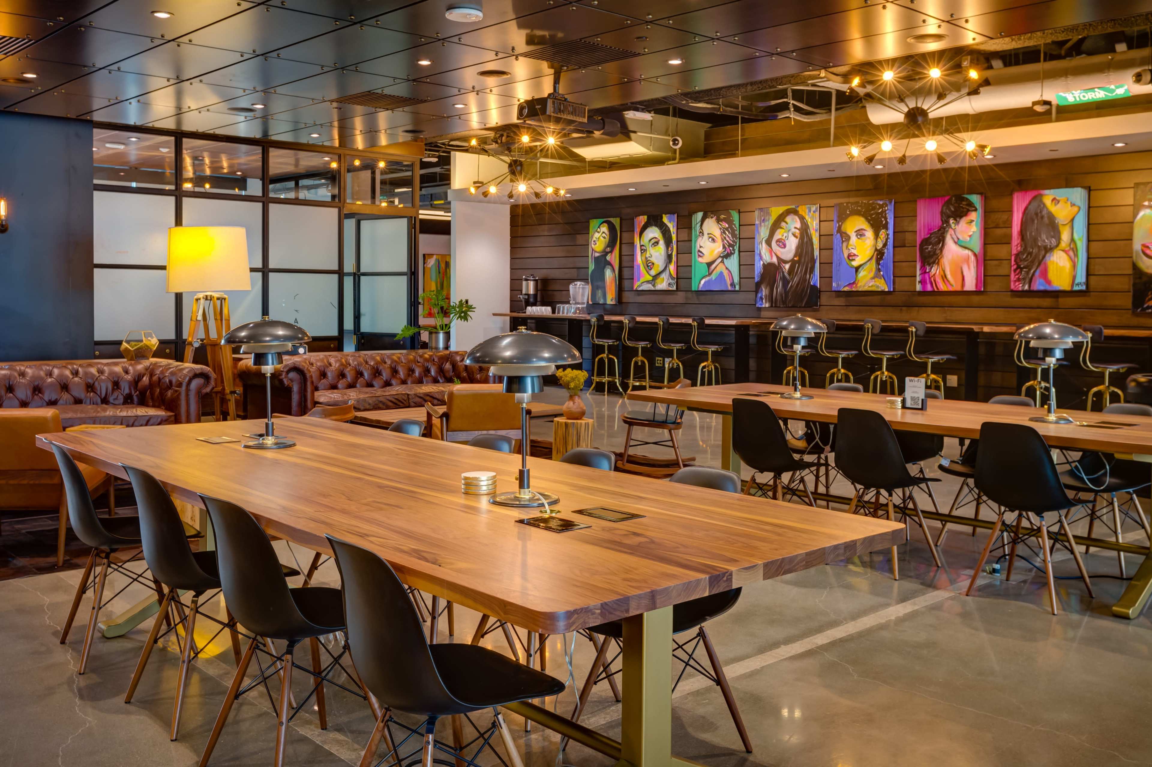 The image shows a modern workspace featuring a large wooden table, black chairs, a leather couch, and colorful artwork displayed on the walls.