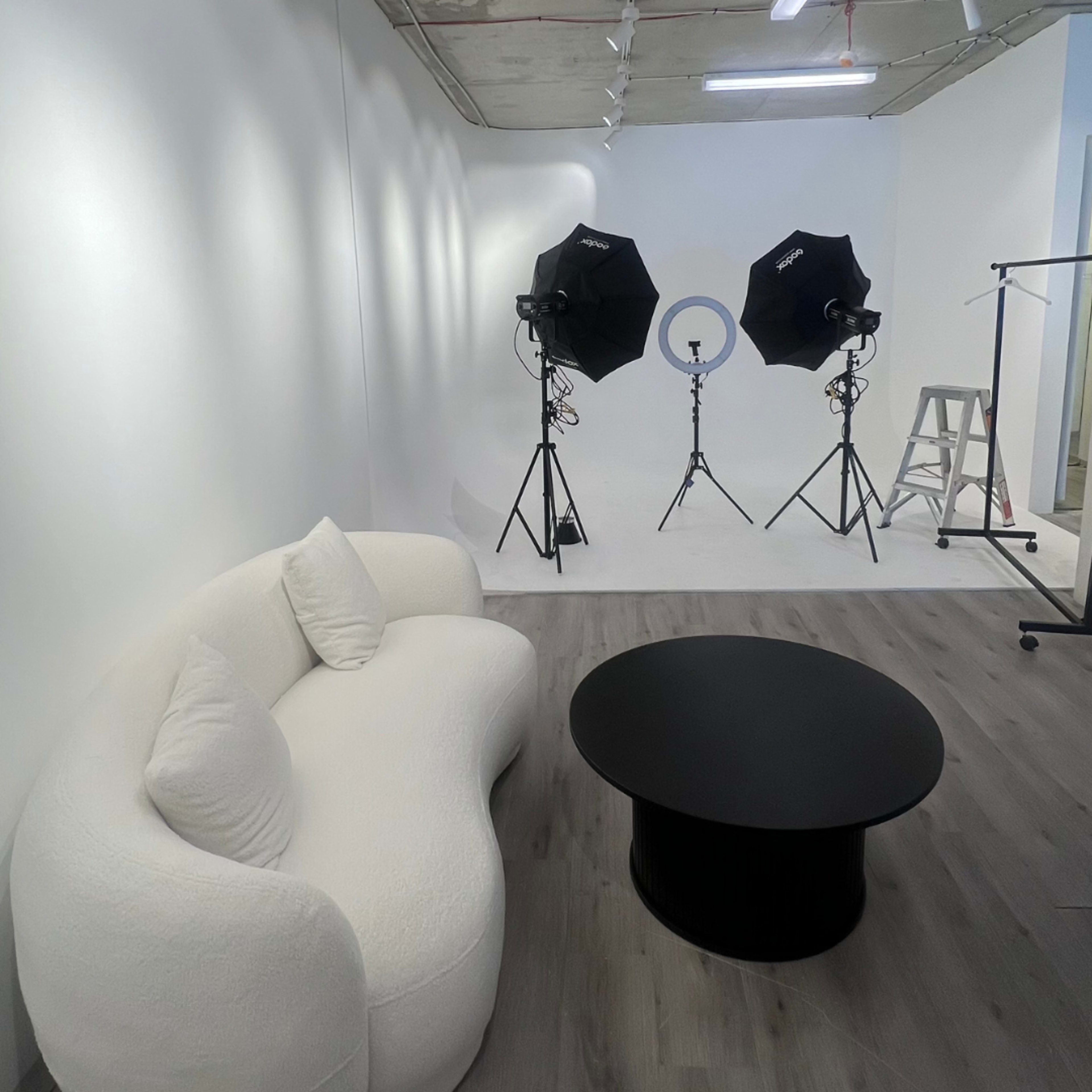 The image shows a photography studio featuring a white curved couch and a black circular coffee table, with two softbox lights positioned on tripods against a white backdrop.