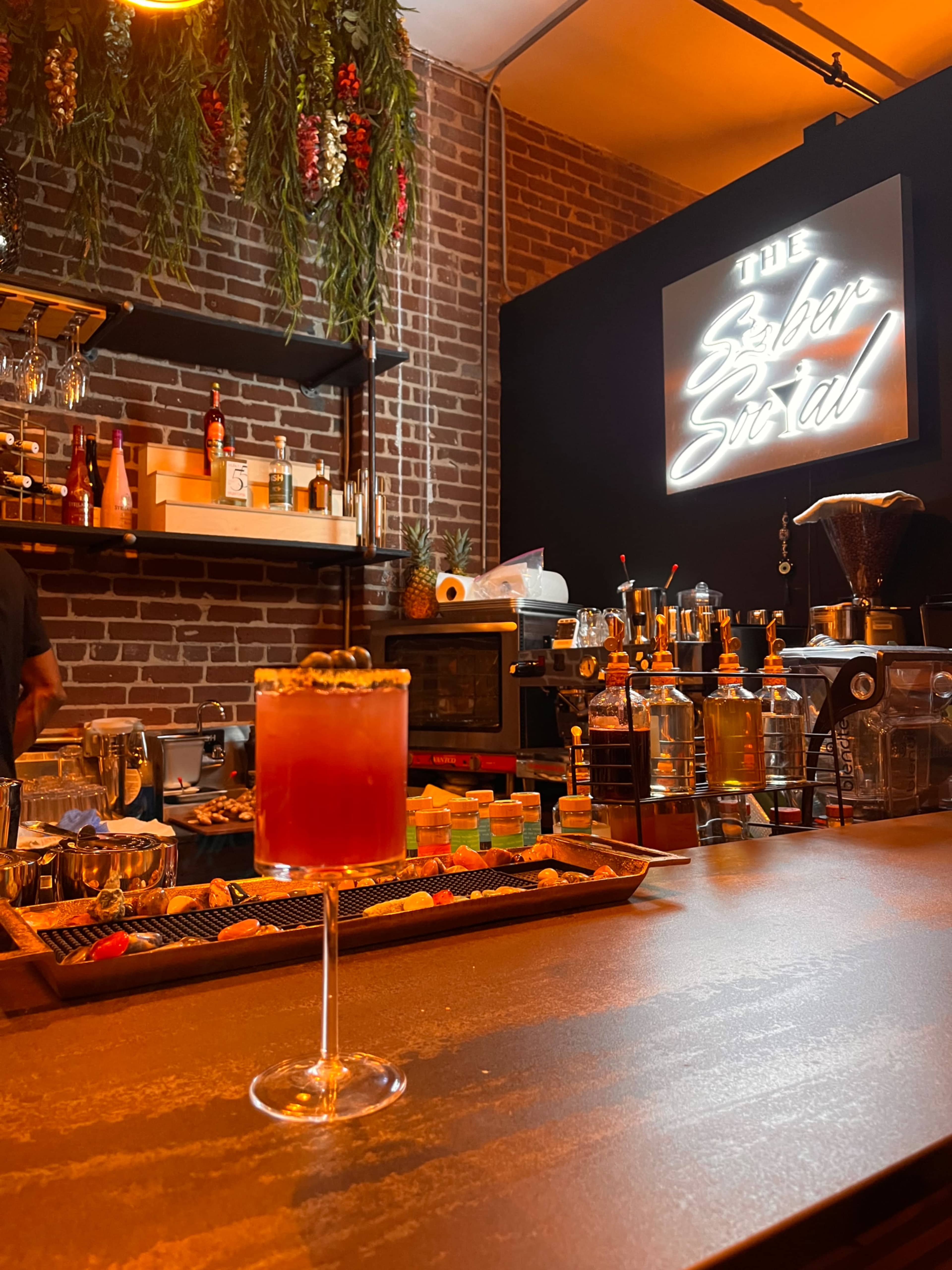 A dimly lit bar scene with a cocktail on the counter in the foreground, and a stylish bar setup featuring shelves of liquor and a neon sign in the background.