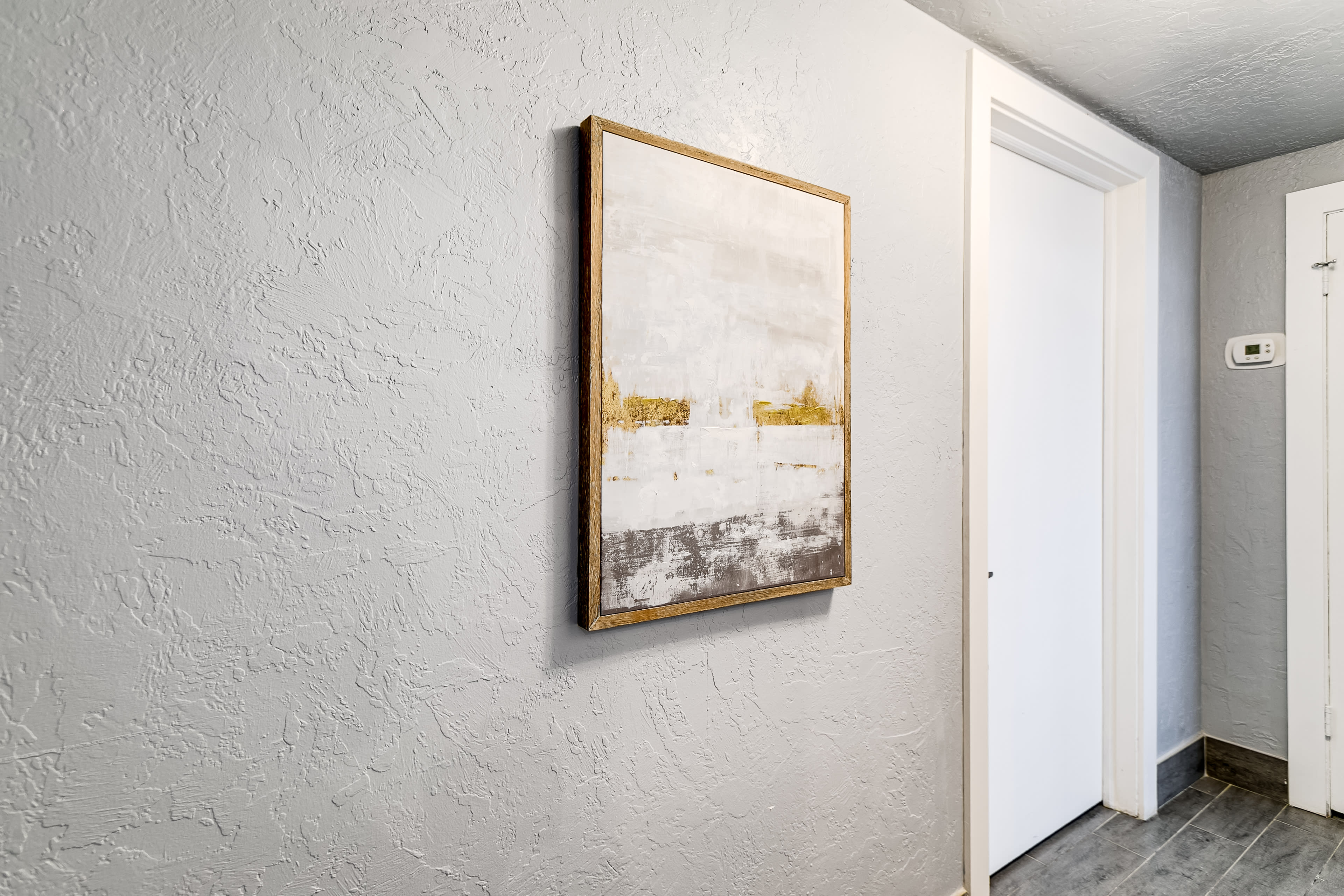 A textured gray wall with a framed artwork hanging on it, next to a white door and a thermostat on the side.