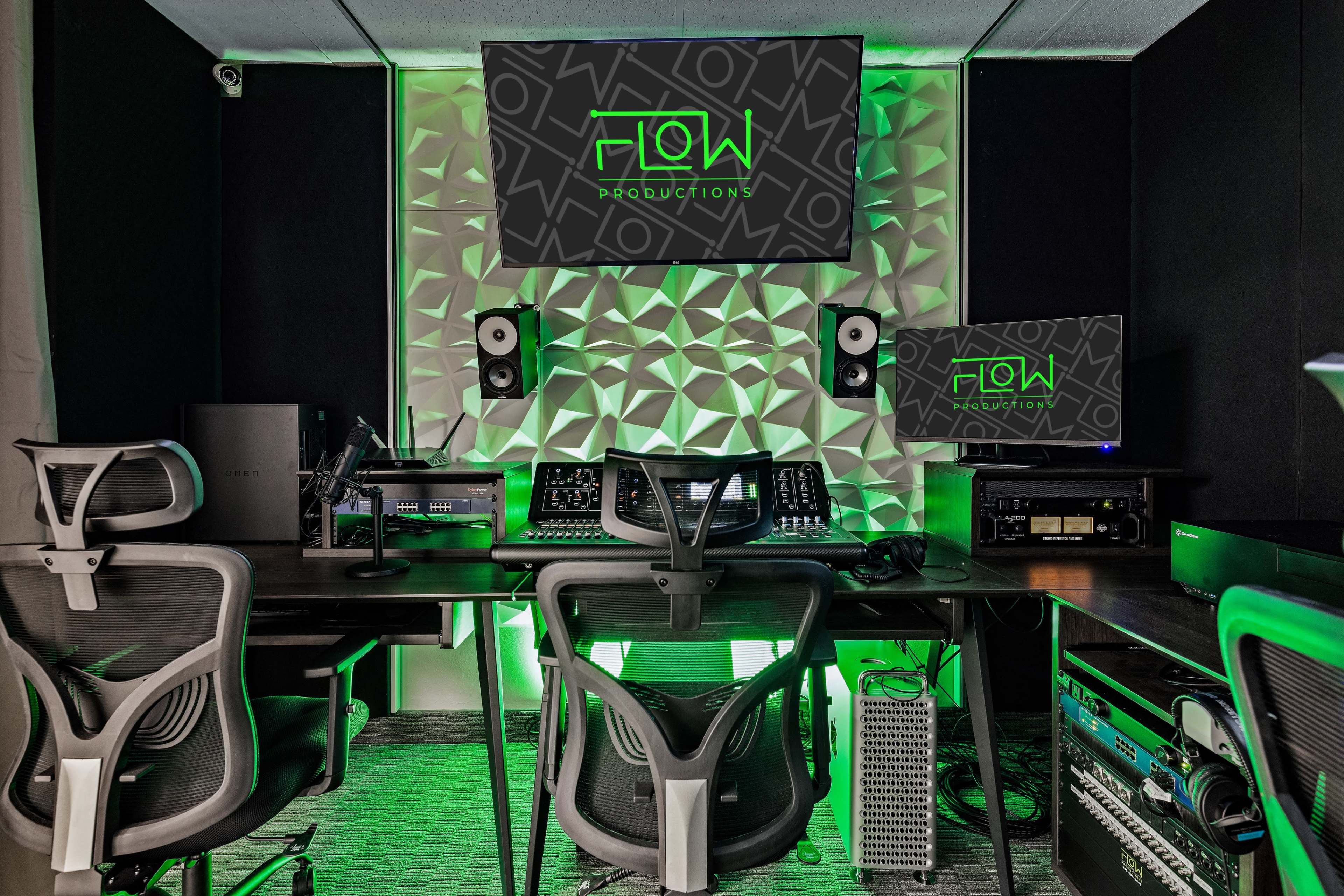 A modern audio production studio featuring desks with ergonomic chairs, two large monitors, and various audio equipment against a textured, illuminated wall.
