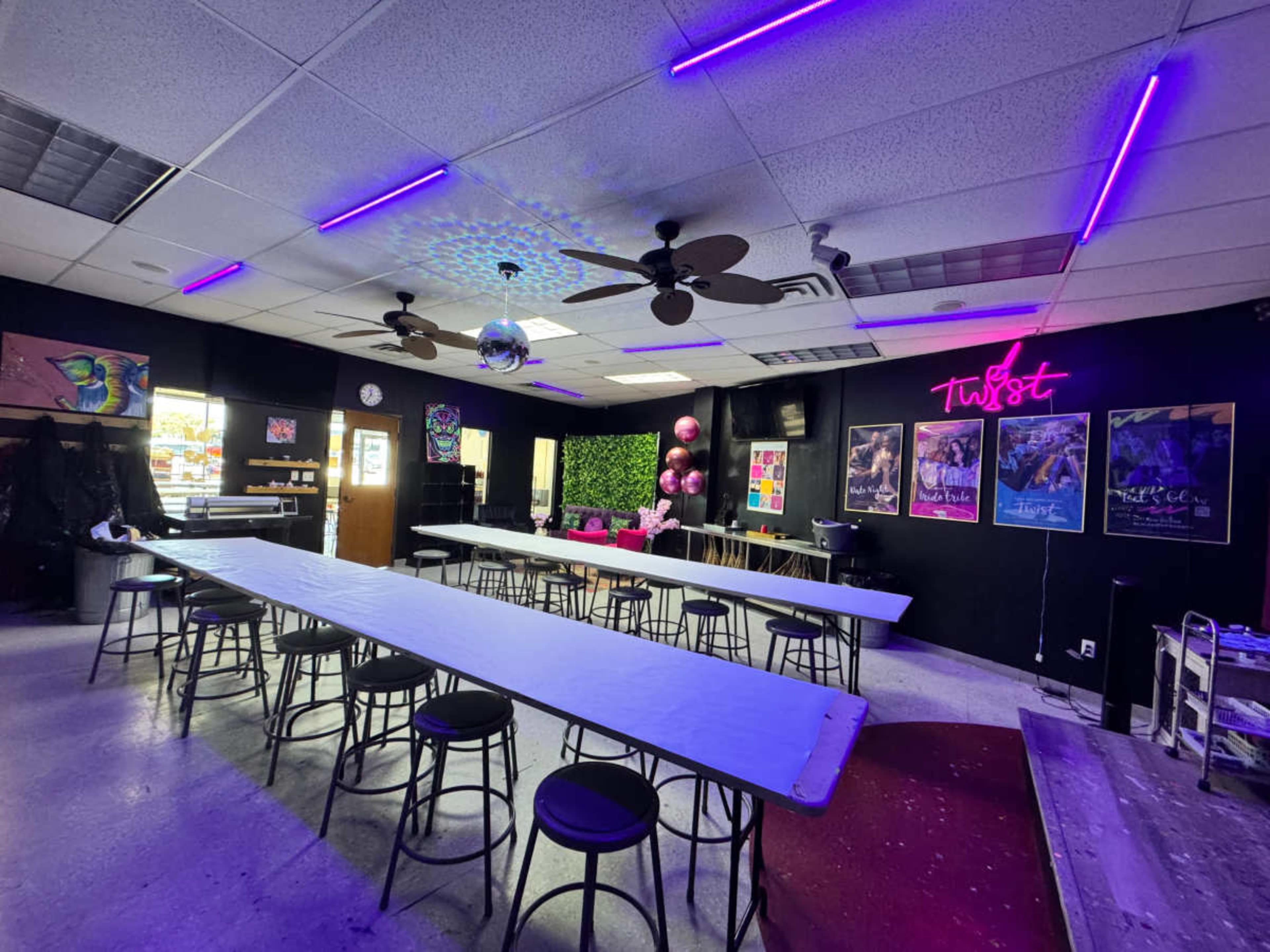 The image shows a brightly colored event space with long tables, black walls, neon lights, and decorations, including a disco ball and pink balloons.
