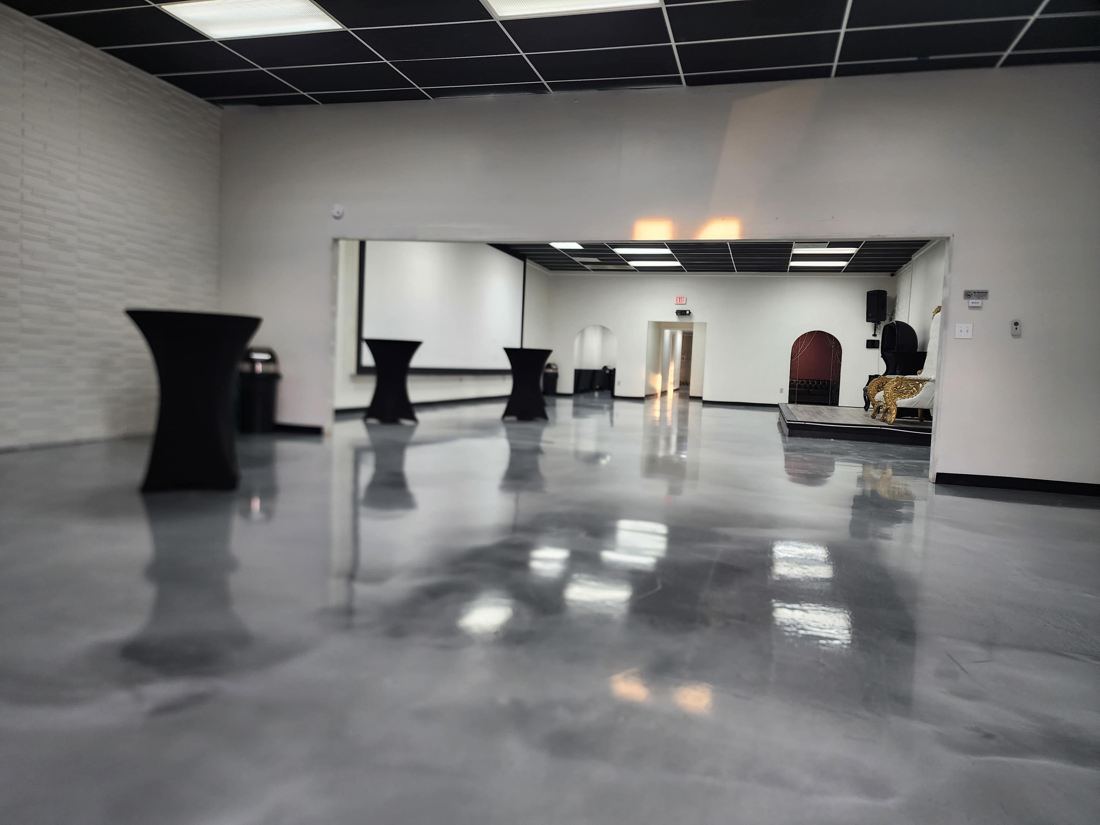 An empty modern event space with polished floors, tall black tables, and an elevated stage at the far end.