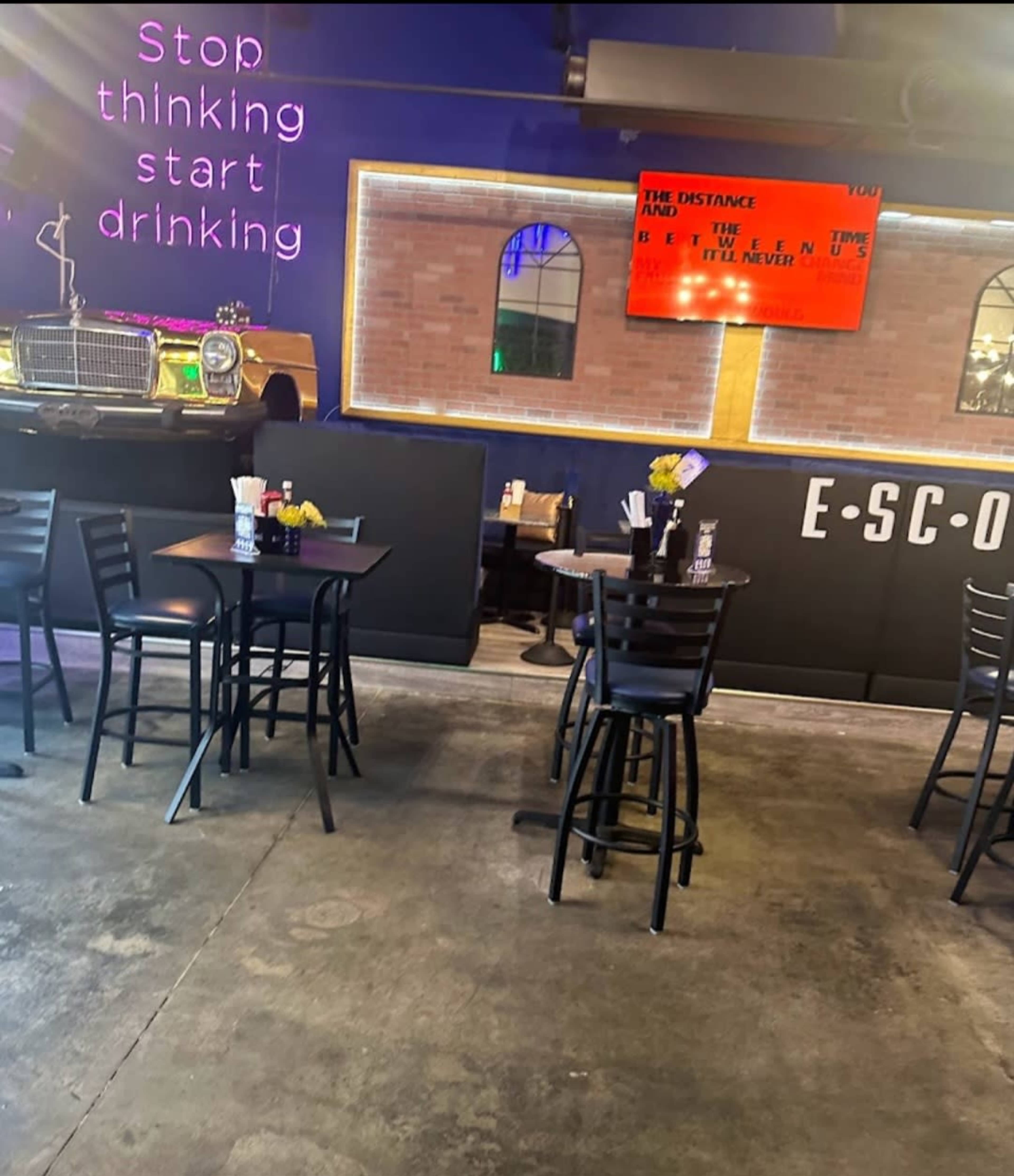 The image shows a restaurant interior with several black tables and chairs, illuminated by neon signs that read "Stop thinking, start drinking," and a digital screen displaying text on a wall.