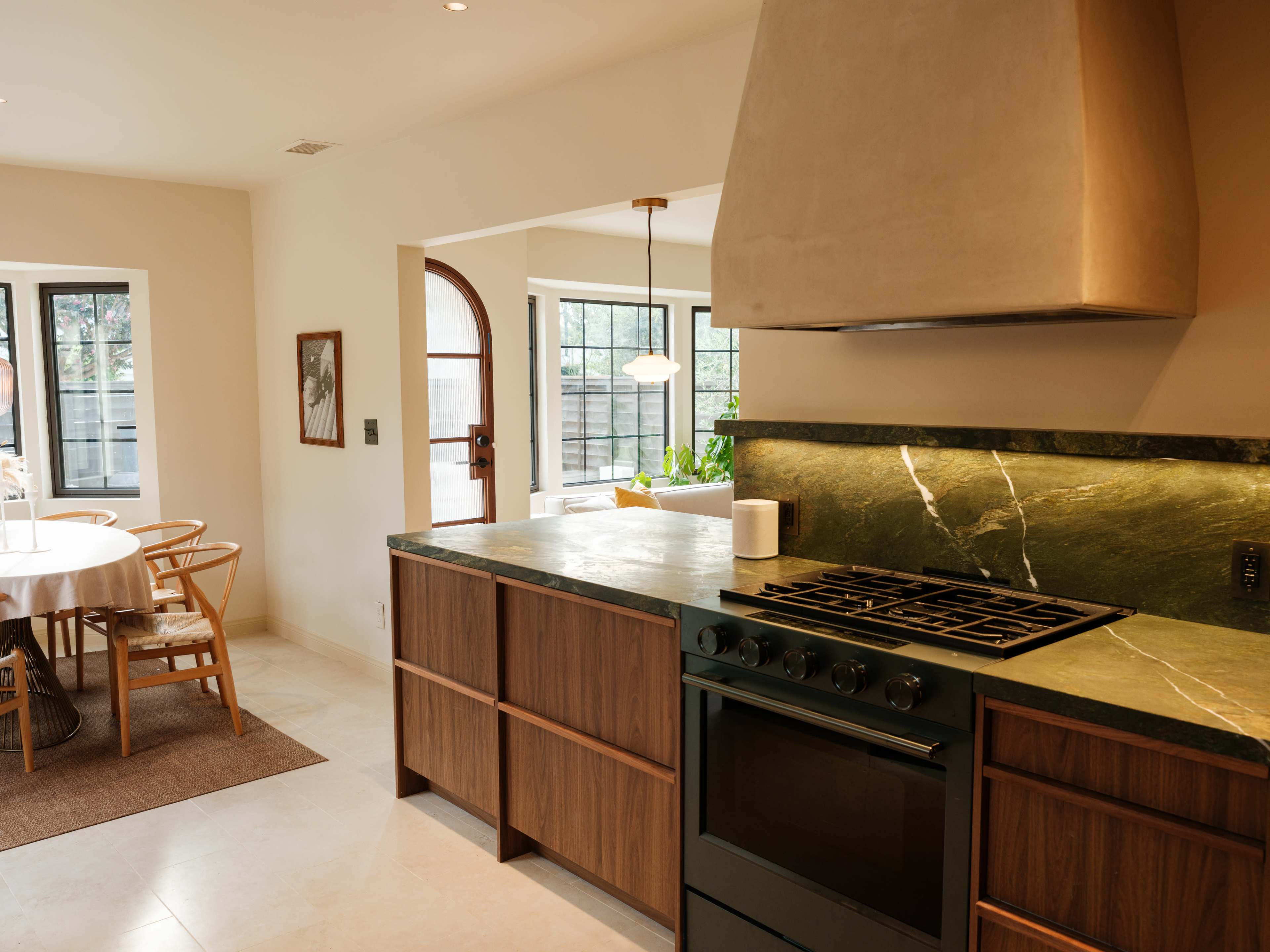 The image shows a modern kitchen with a green stone countertop, wooden cabinetry, and a gas stove, connected to a dining area with a round table and chairs.