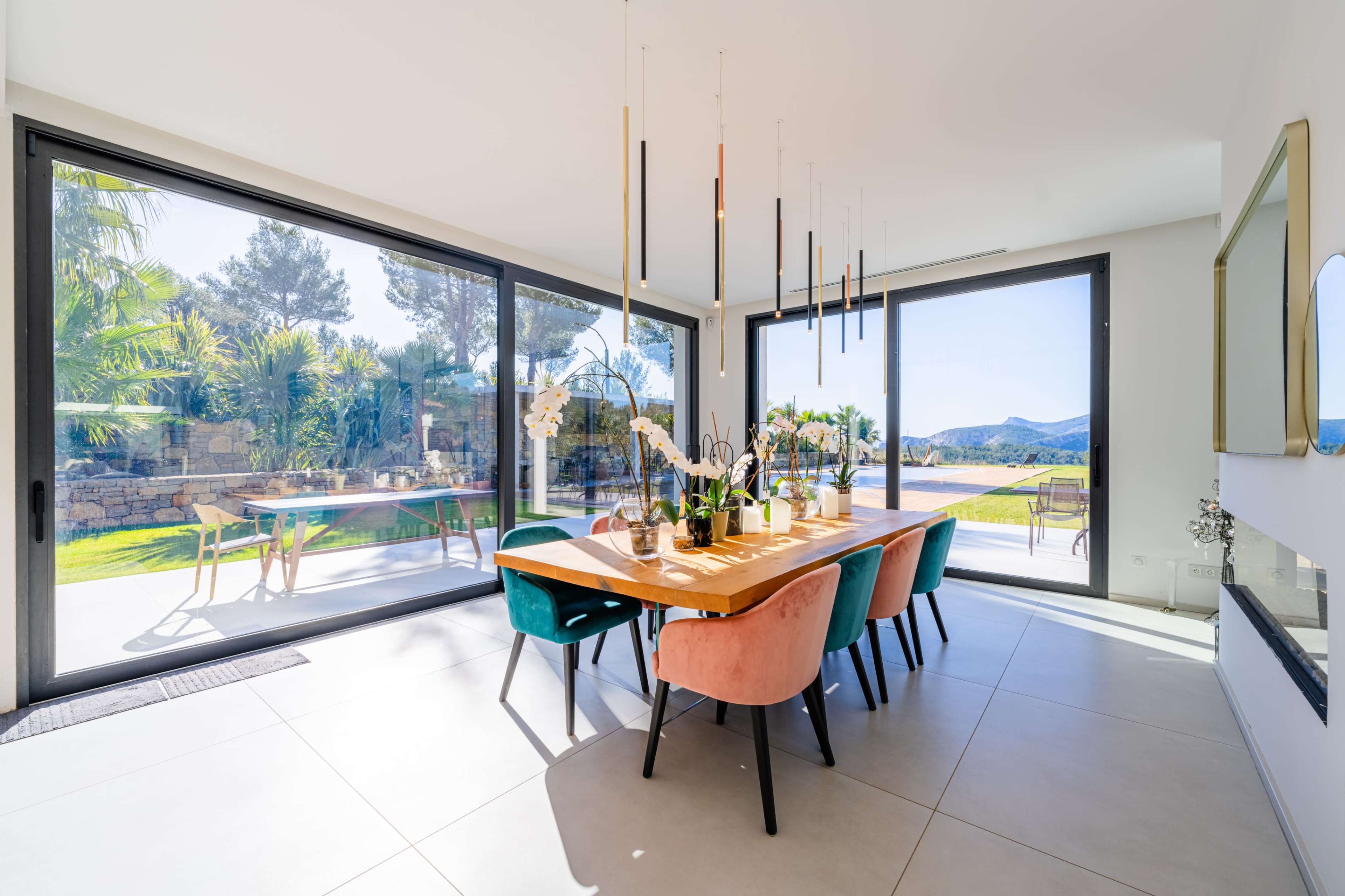 A spacious dining area features a wooden table surrounded by colorful chairs, with panoramic windows showcasing a landscaped outdoor view.