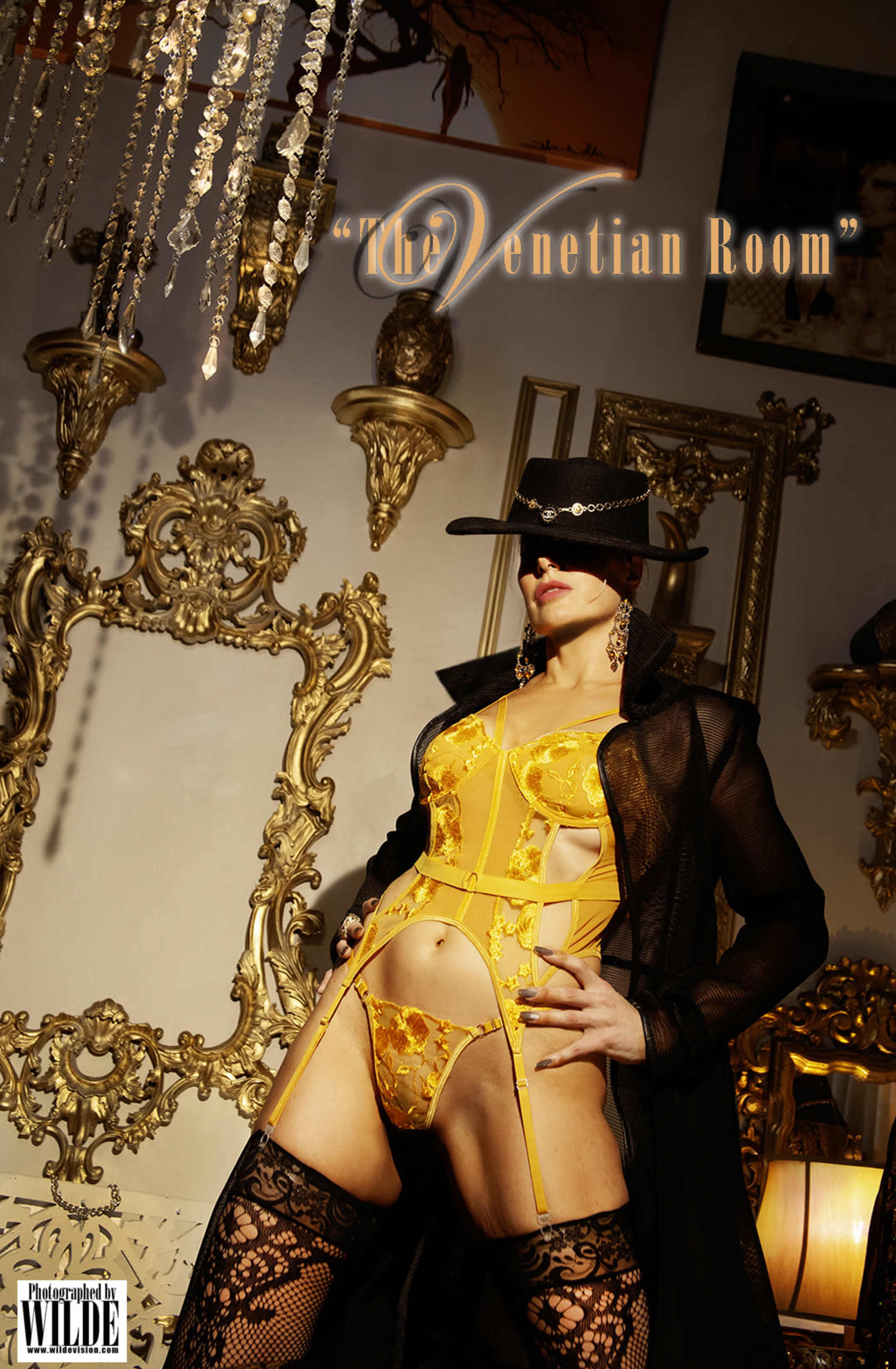 A model poses in a yellow lingerie set and black coat against an ornate backdrop featuring gold-framed mirrors and intricate decor.