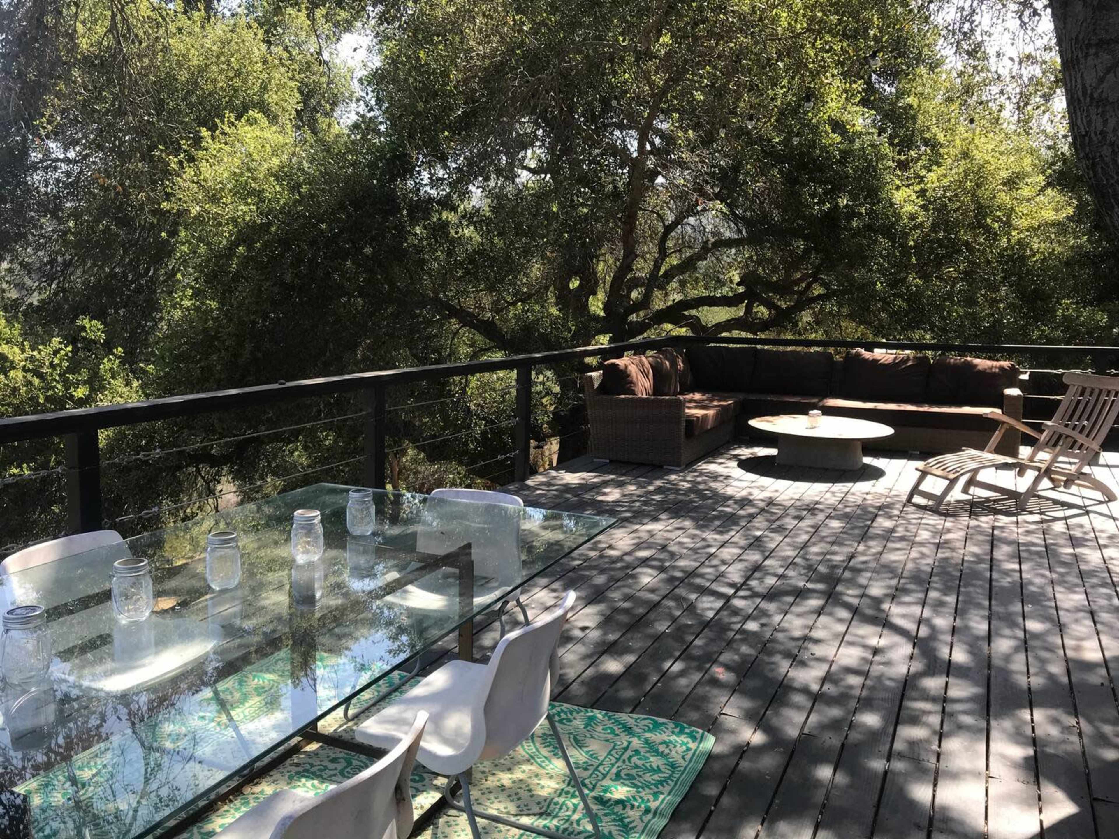 A wooden deck features a glass dining table with chairs, a circular fire pit, and a seating area surrounded by lush greenery.