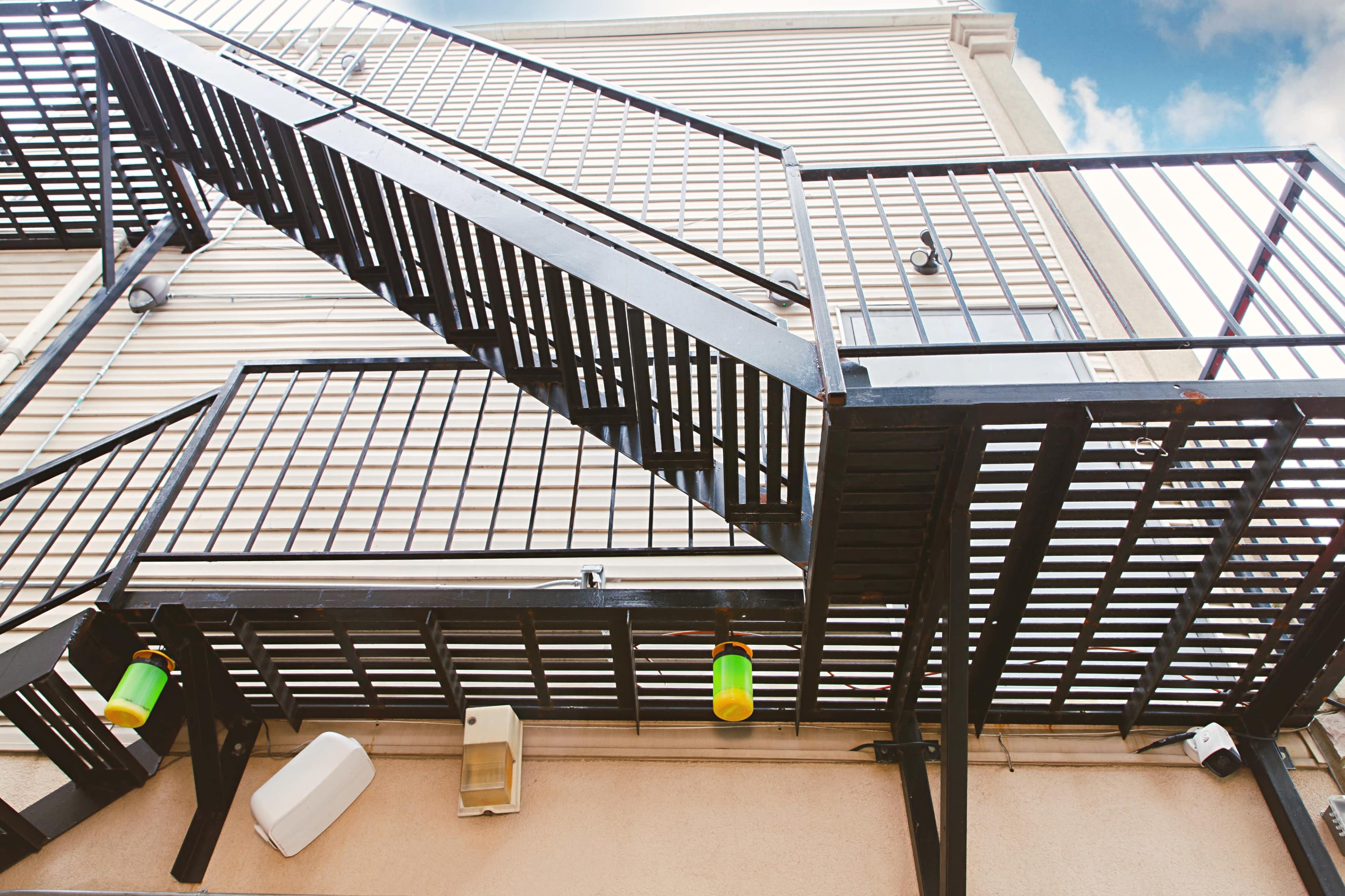 The image shows a black metal fire escape staircase ascending alongside a light-colored building.