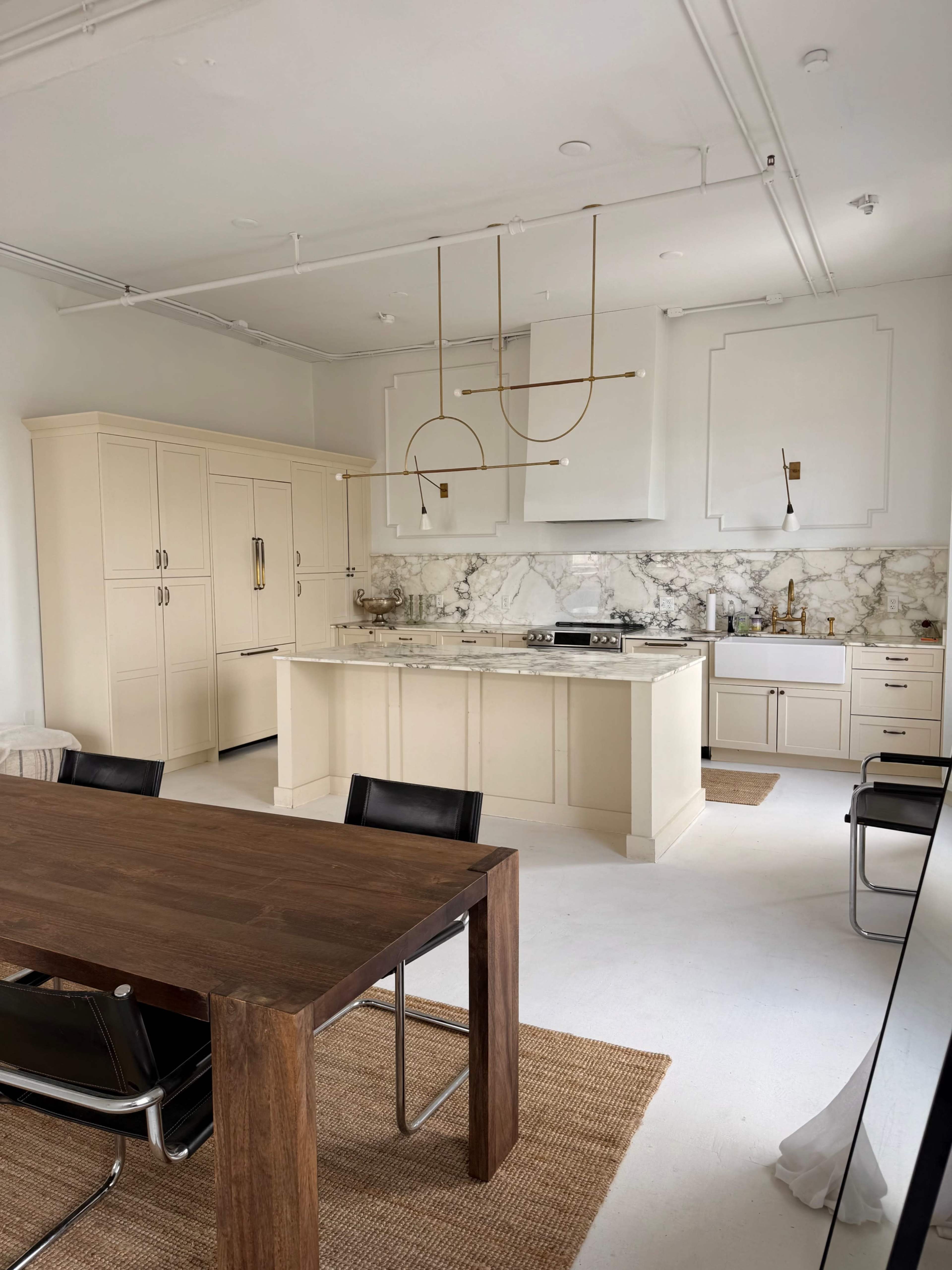 The image shows a modern kitchen with cream-colored cabinetry, a marble backsplash, and a wooden dining table surrounded by black chairs.