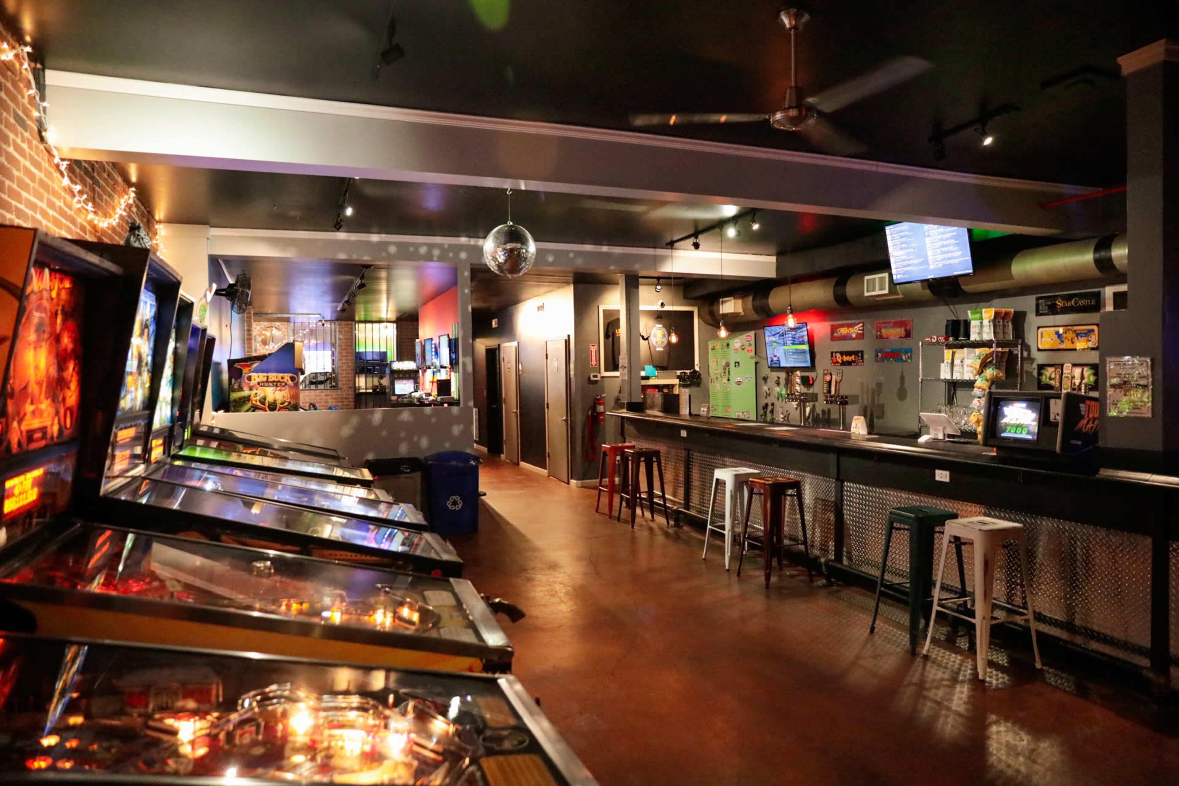 A dimly lit bar with several pinball machines on the left and a bar area with stools and a display screen on the right.