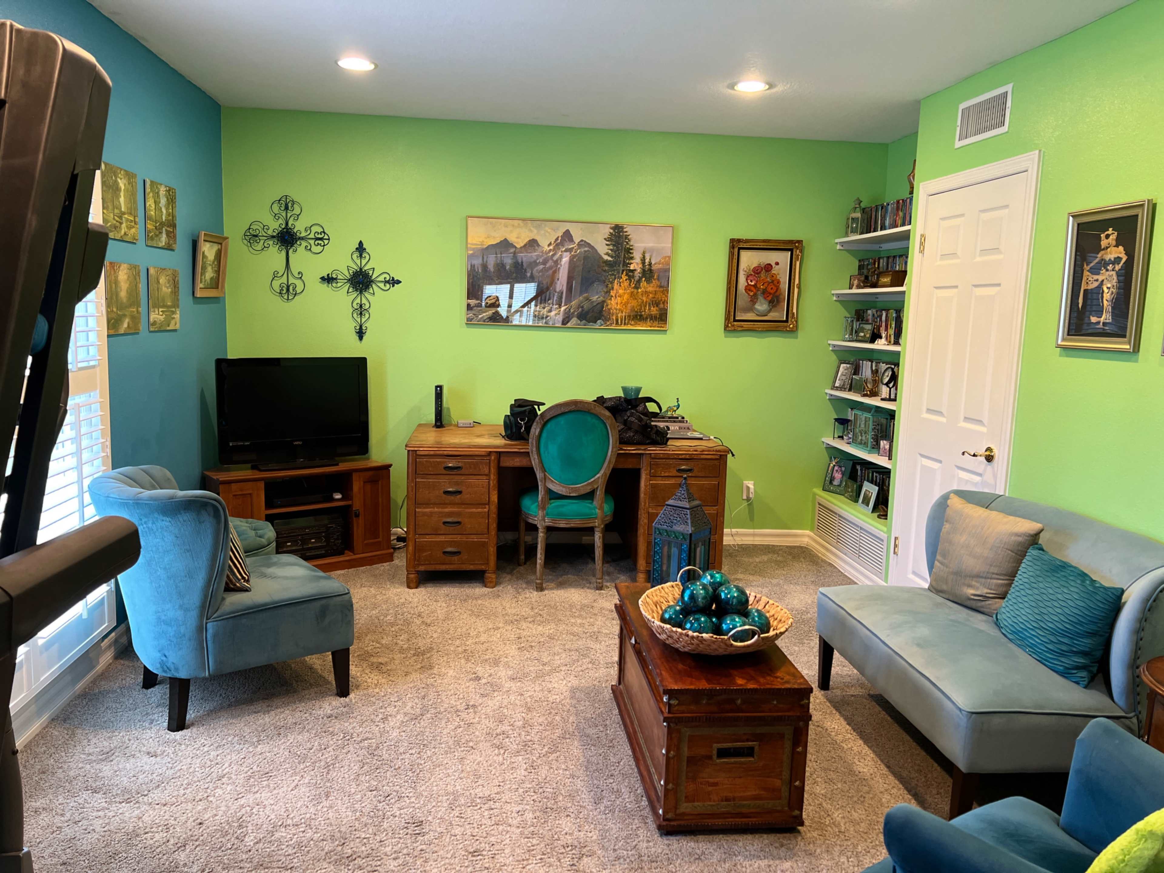 The room features a green and blue color scheme with a desk, chairs, a television, and various decorations on the walls and shelves.