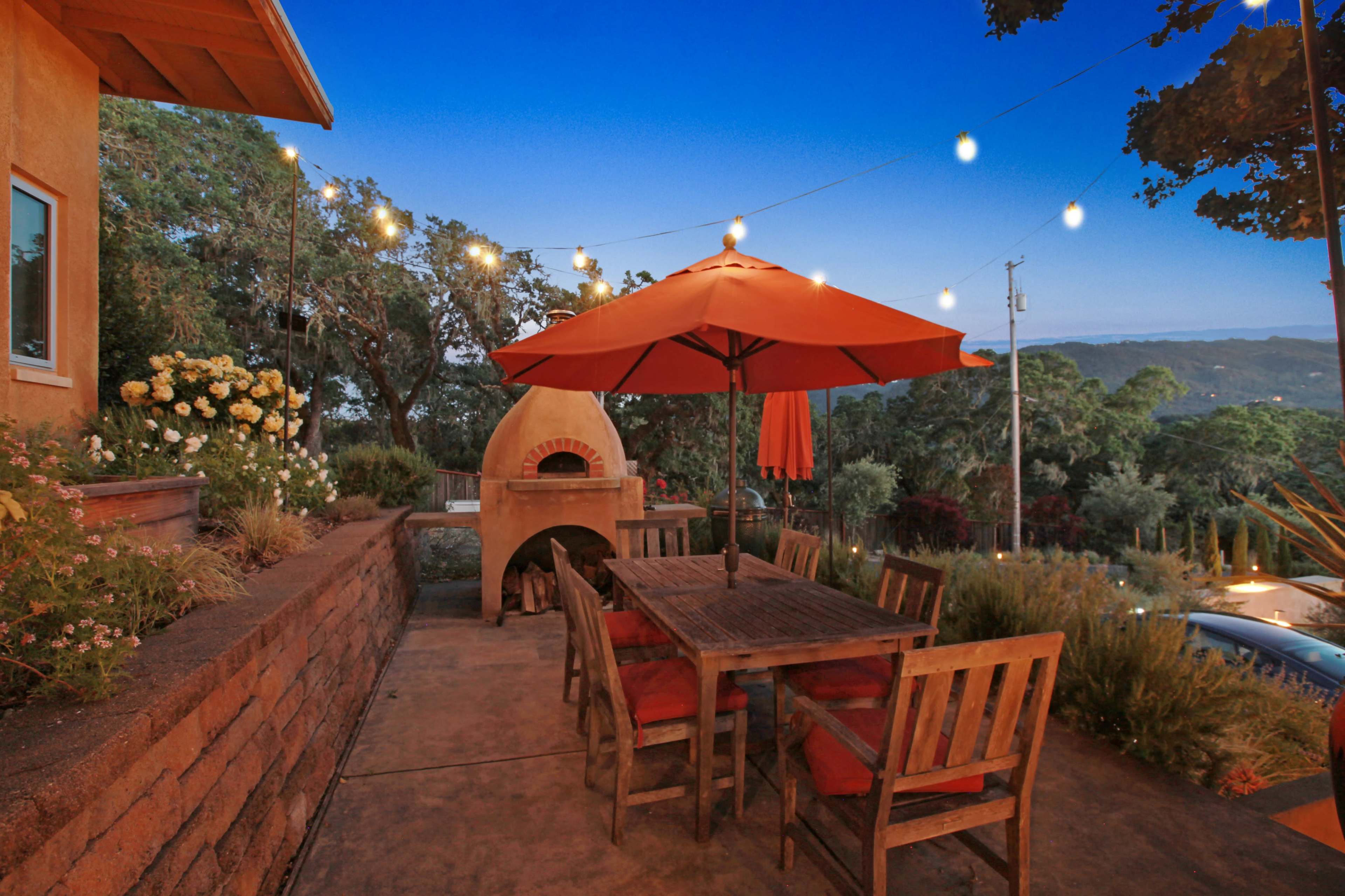 A wooden dining table with chairs is set under an orange umbrella beside a stone pizza oven, illuminated by string lights against a twilight sky.