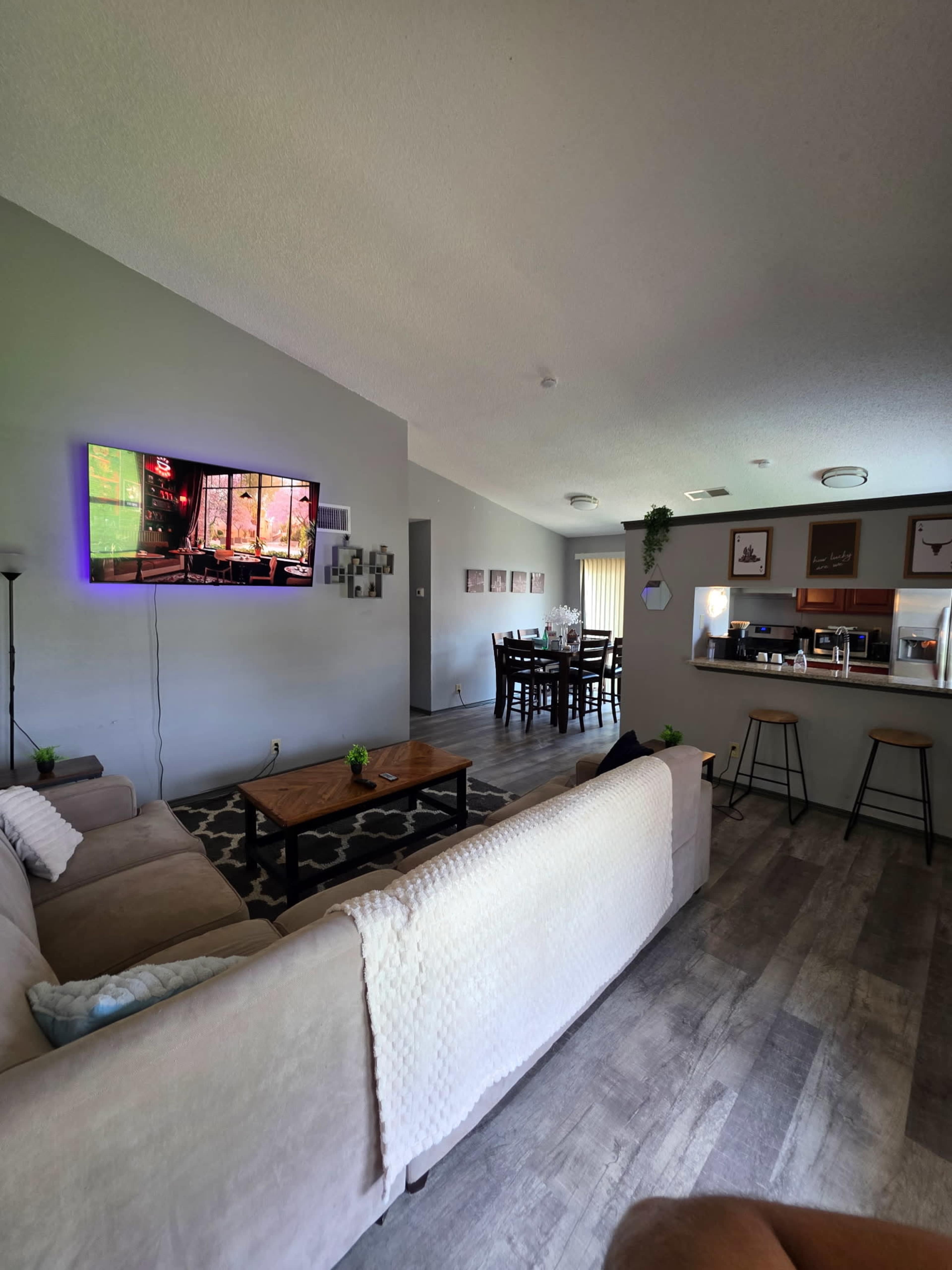 The image shows a modern living room with a large television mounted on the wall, a beige couch, a coffee table, and a dining area visible in the background.
