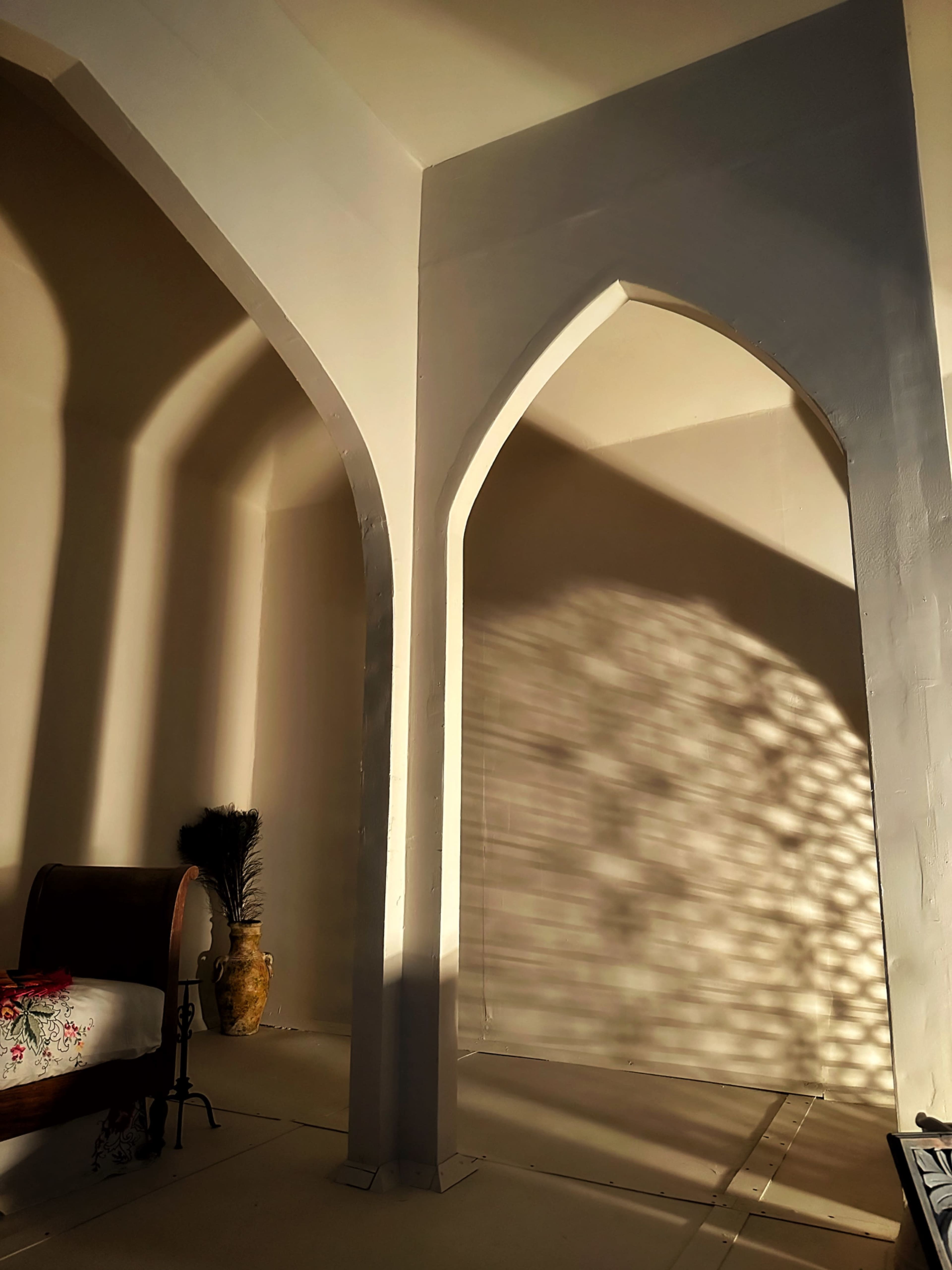 The image shows a corner of a room with arched walls casting shadows, a wooden bench, and a vase.