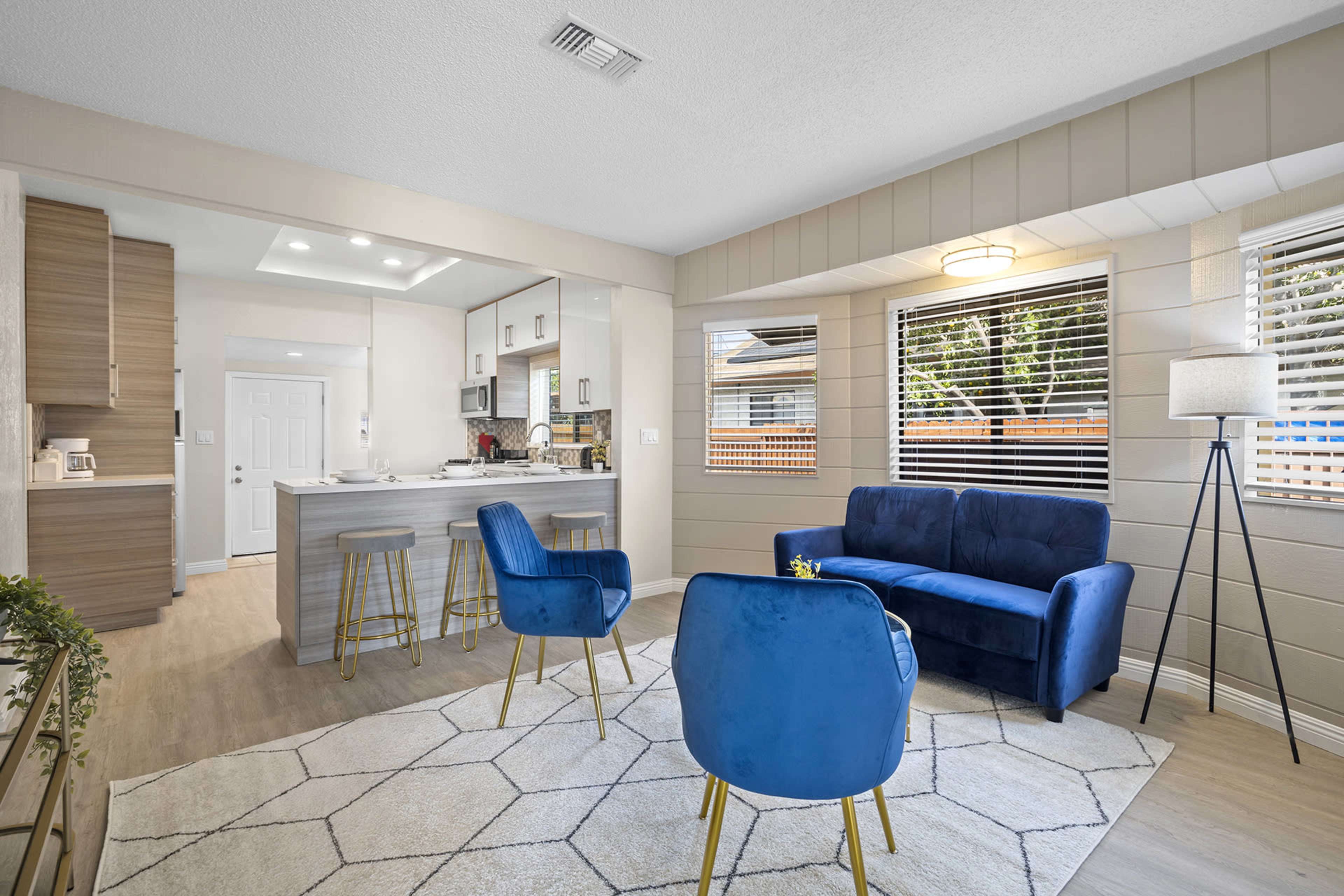 A modern living space featuring a blue sofa, two blue chairs, a geometric rug, and a kitchen area with a breakfast bar in the background.