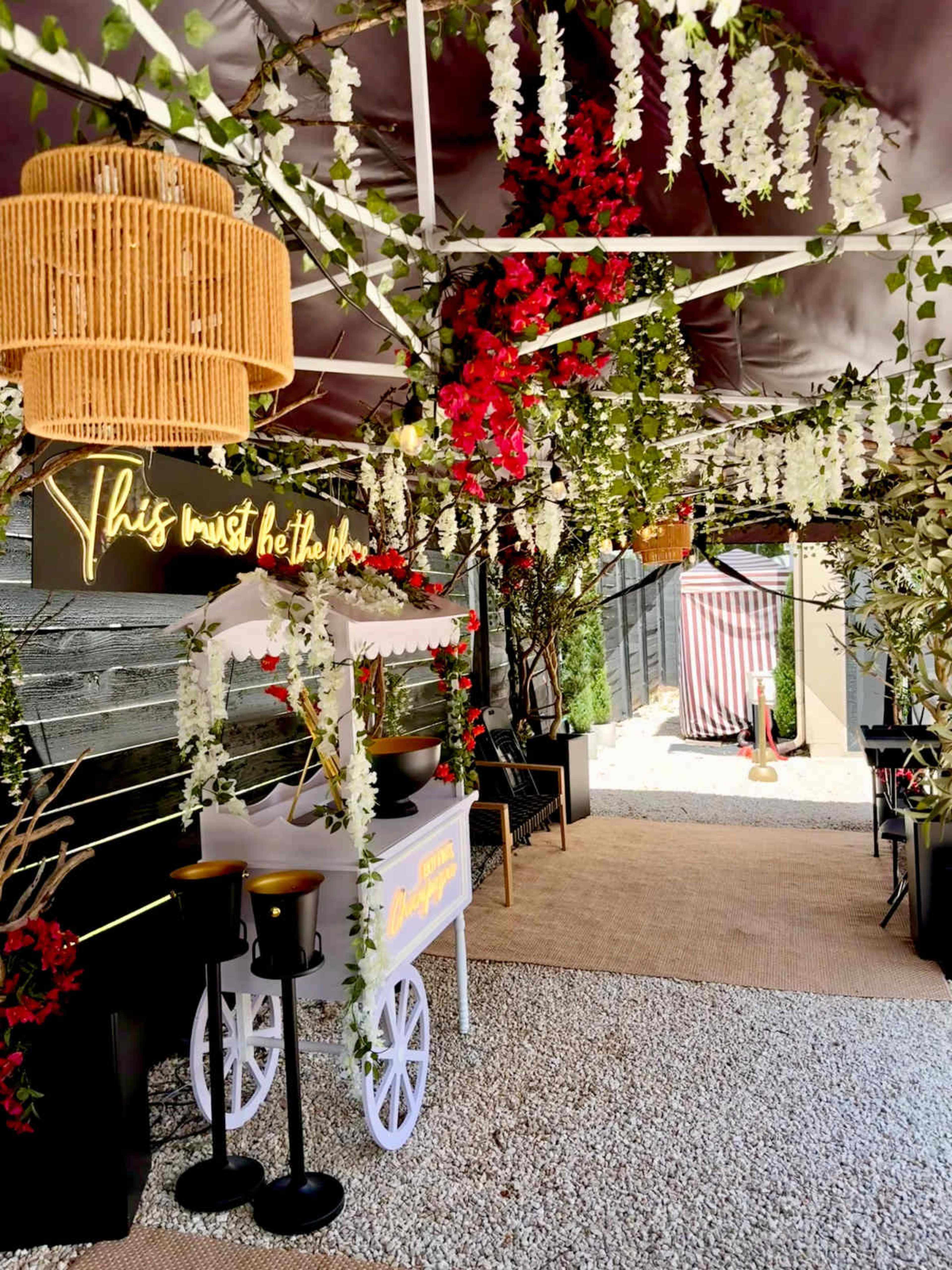 The image shows a decorated outdoor pathway adorned with hanging flowers, a warm light fixture, and a whimsical sign.